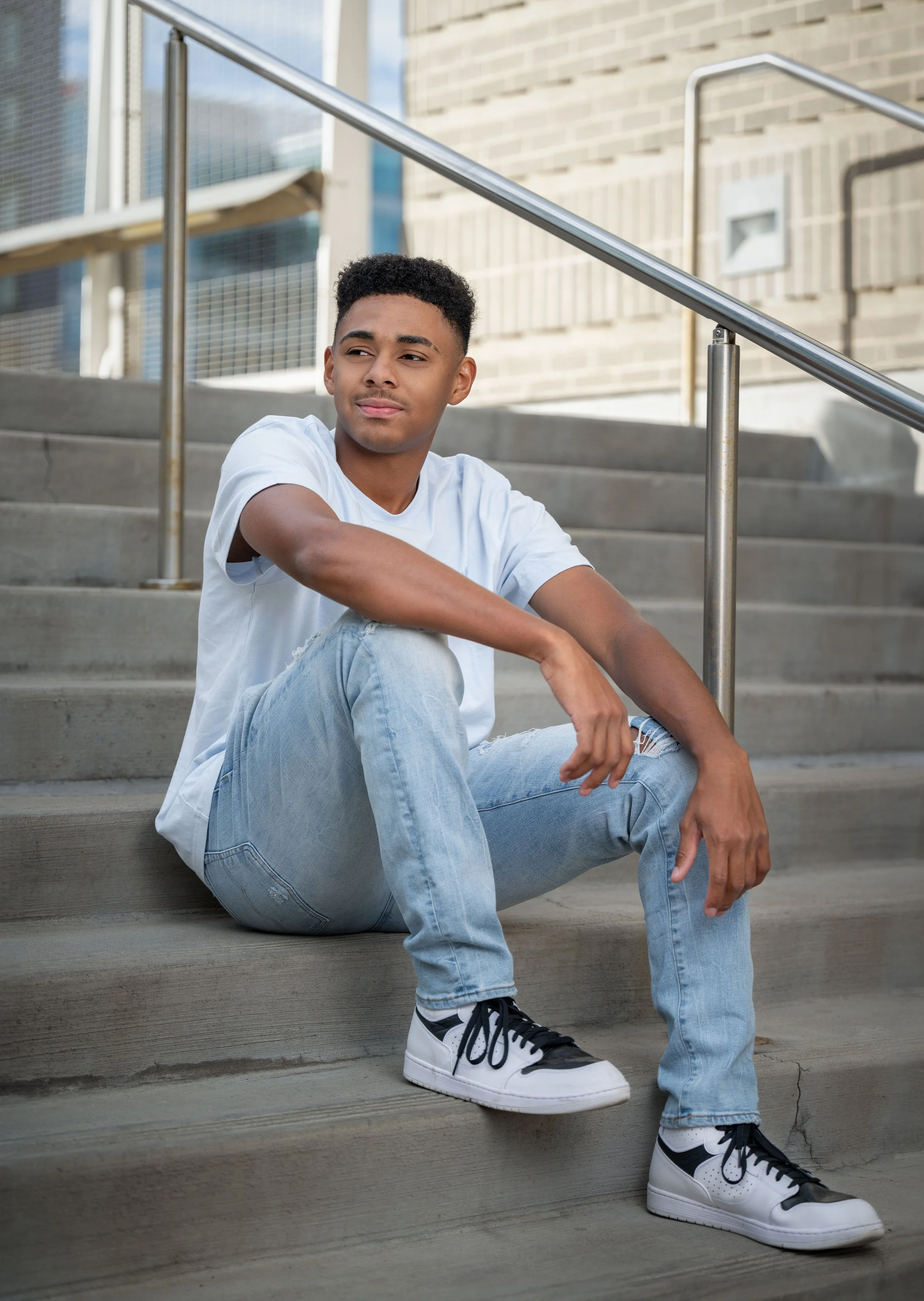 A young man sitting on outdoor concrete steps beside a metal railing, wearing a white T-shirt, light blue jeans, and white and black sneakers, with a contemplative expression.