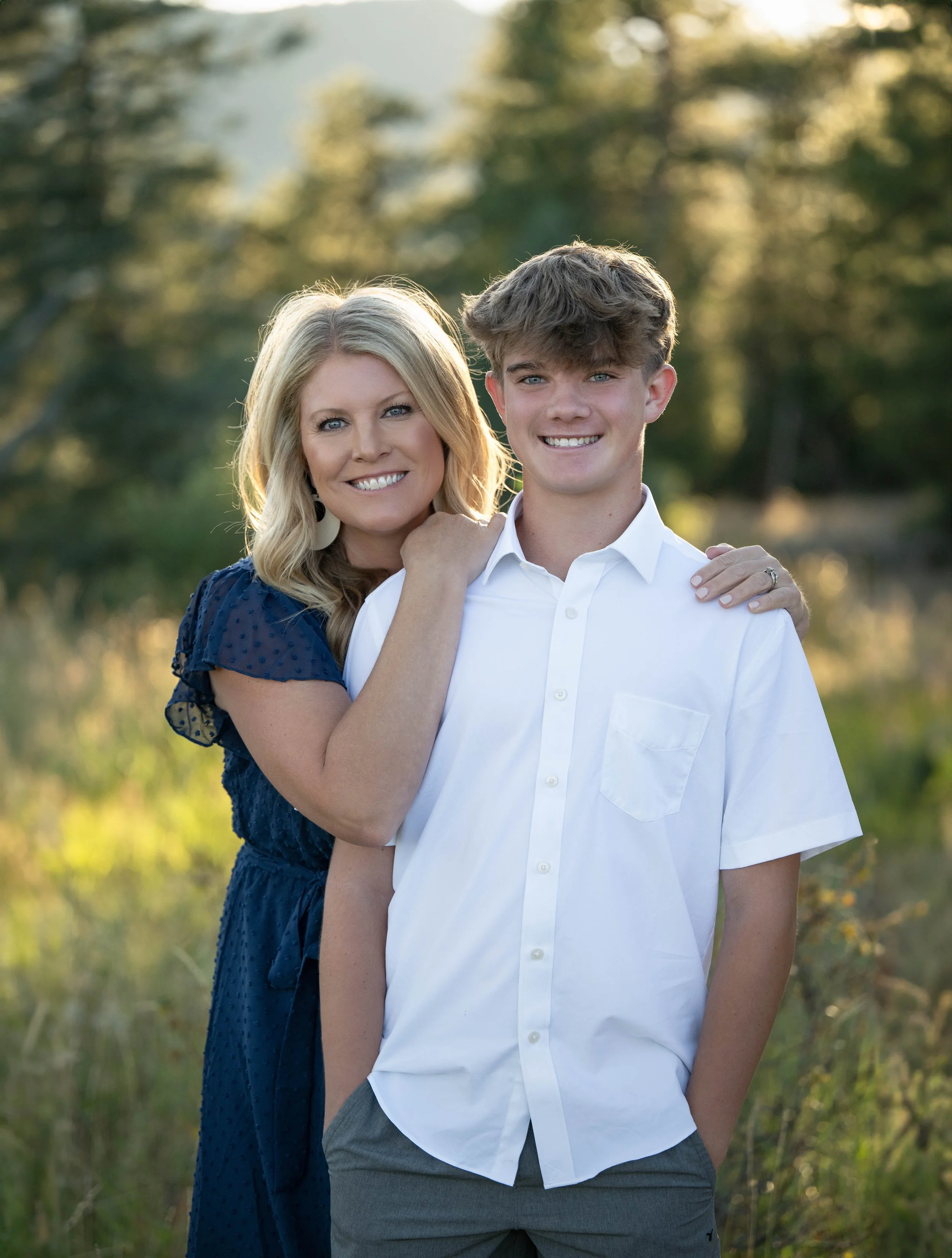 A woman and teenage boy standing outdoors in a nature setting, smiling at the camera, with the woman resting her hand on the boy's shoulder.