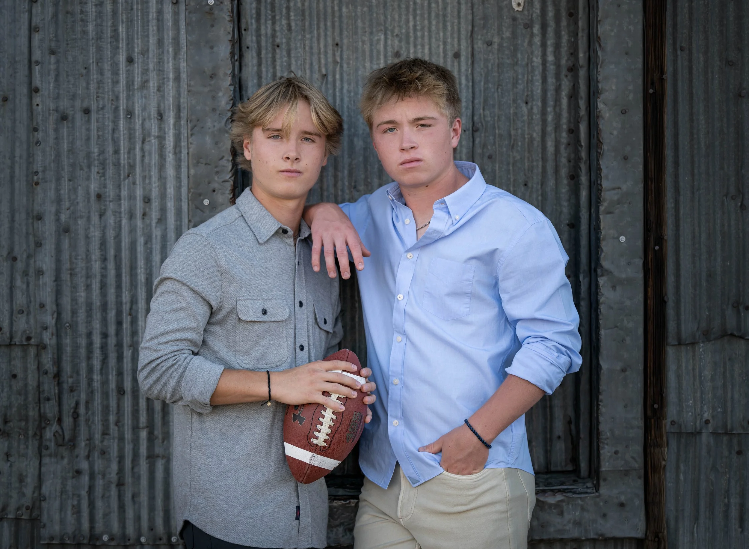 Two teenage boys standing close together against a weathered metal wall, one holding a football, both looking serious.