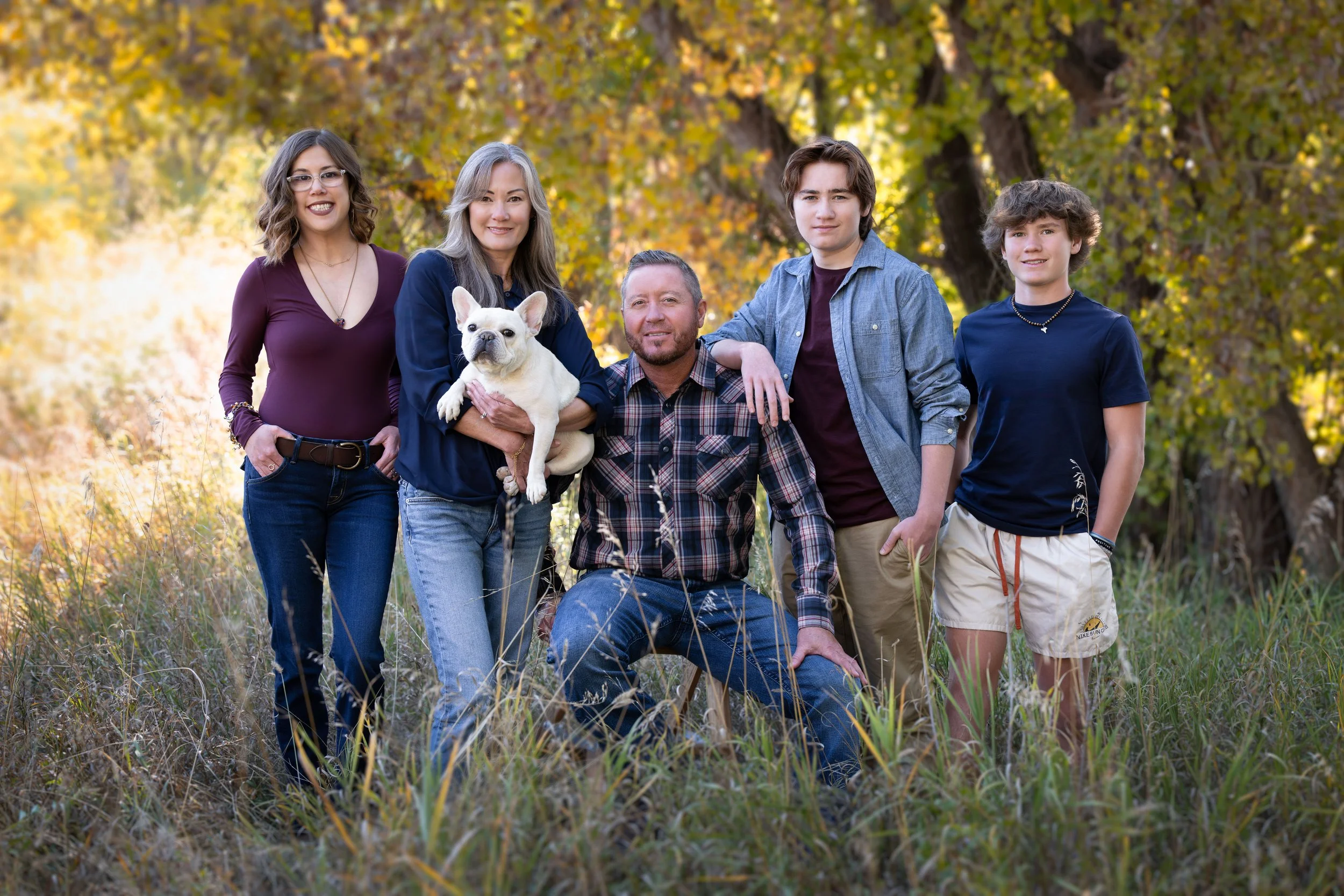 A family of six including three women, two men, and a French bulldog, standing outdoors in a grassy area with trees featuring yellow and orange autumn leaves in the background.