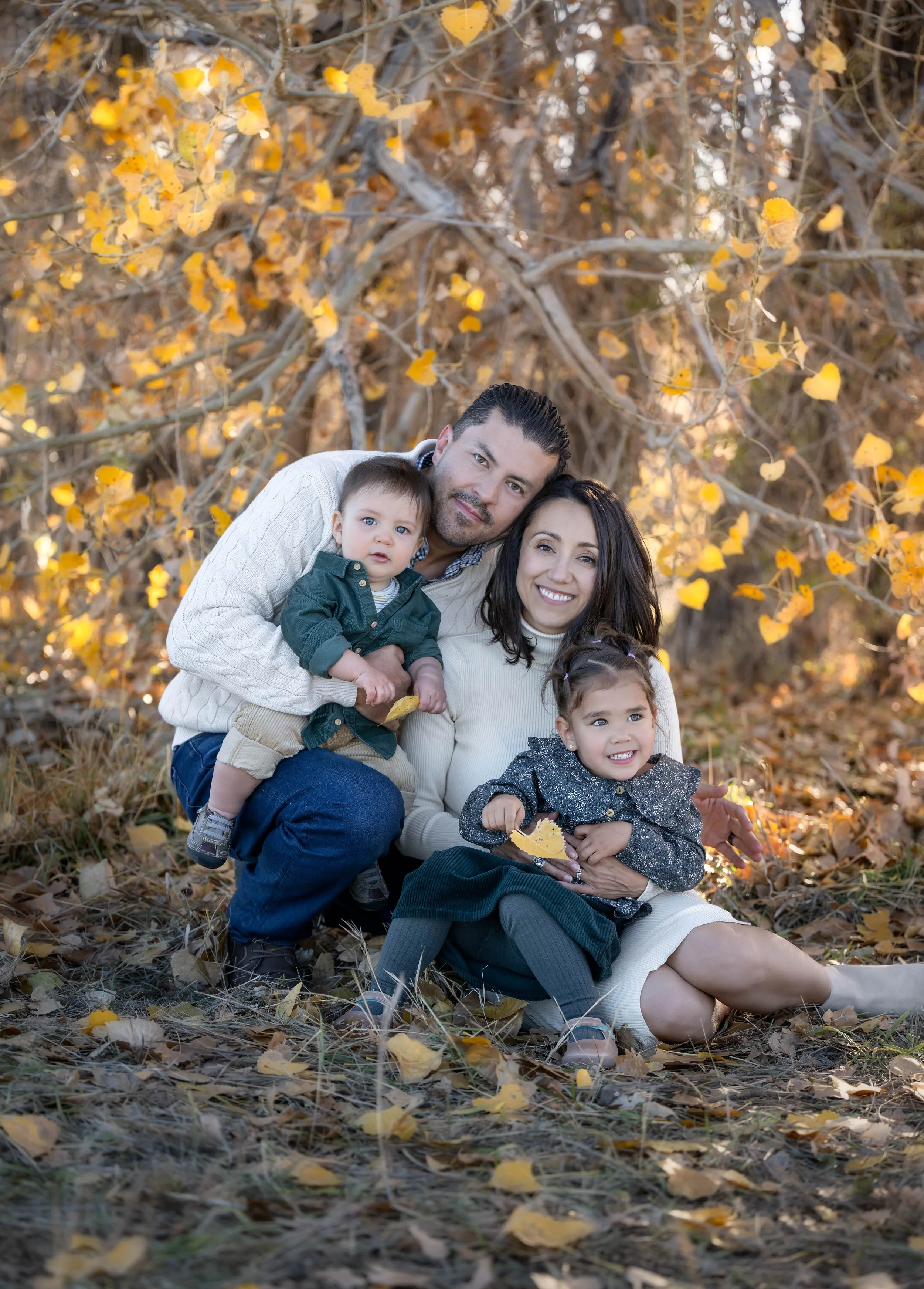 Family of four sitting outdoors on autumn leaves, surrounded by trees with yellow-orange foliage, smiling for the camera.