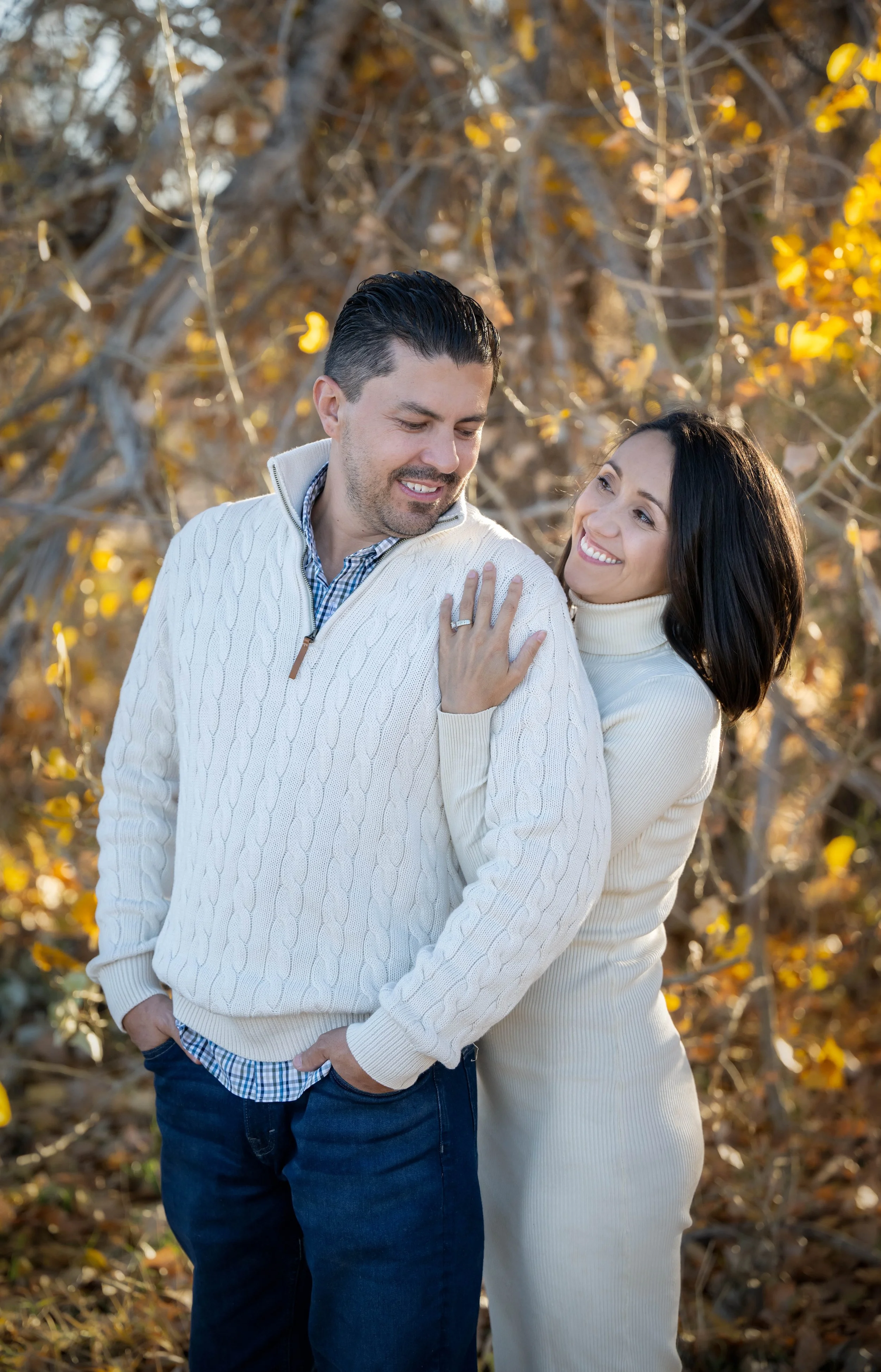 A happy couple outdoors in autumn, with fall foliage in the background, smiling and looking at each other. The woman has her hand on the man's shoulder, showing a ring.