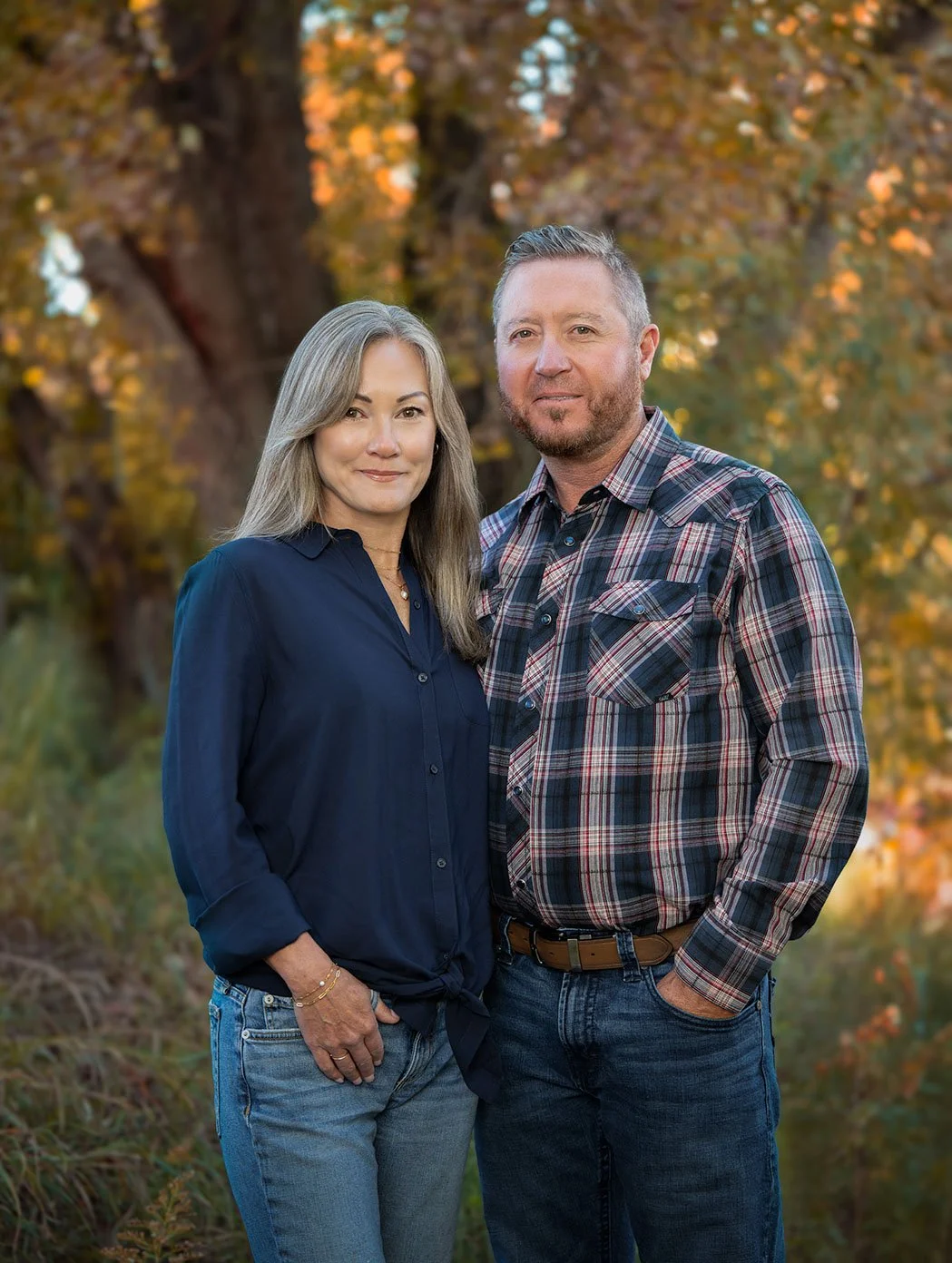 A middle-aged woman and man standing together outdoors with autumn trees in the background, both smiling softly.