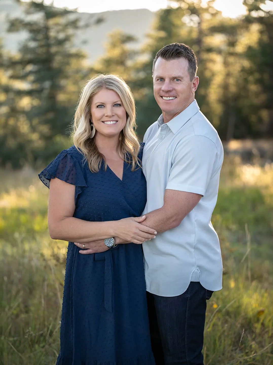 A smiling couple standing outdoors during golden hour, embracing each other with a blurred natural background of trees and water.
