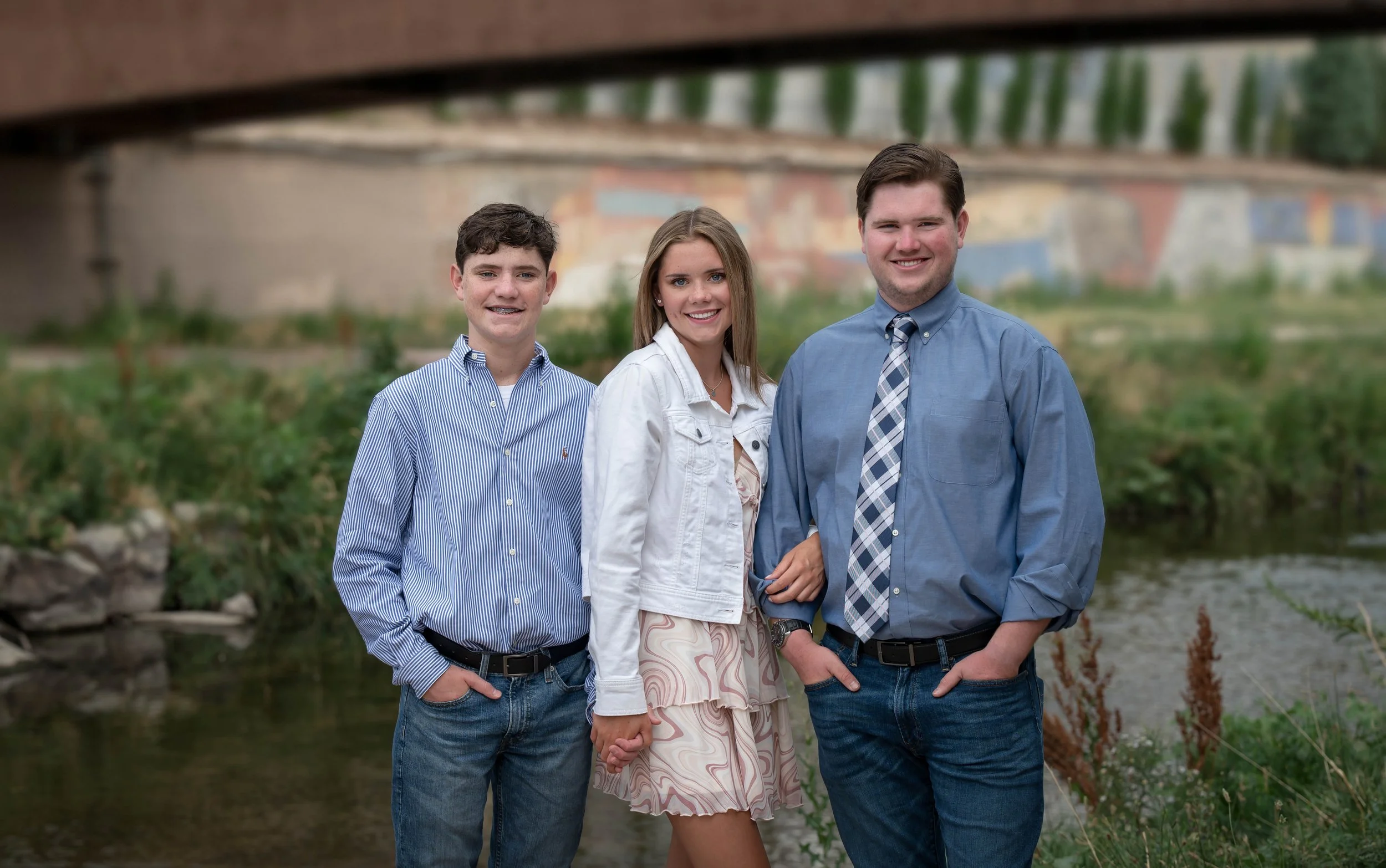 Three young adults, two men and a woman, standing outdoors near a creek with a bridge in the background, smiling at the camera.