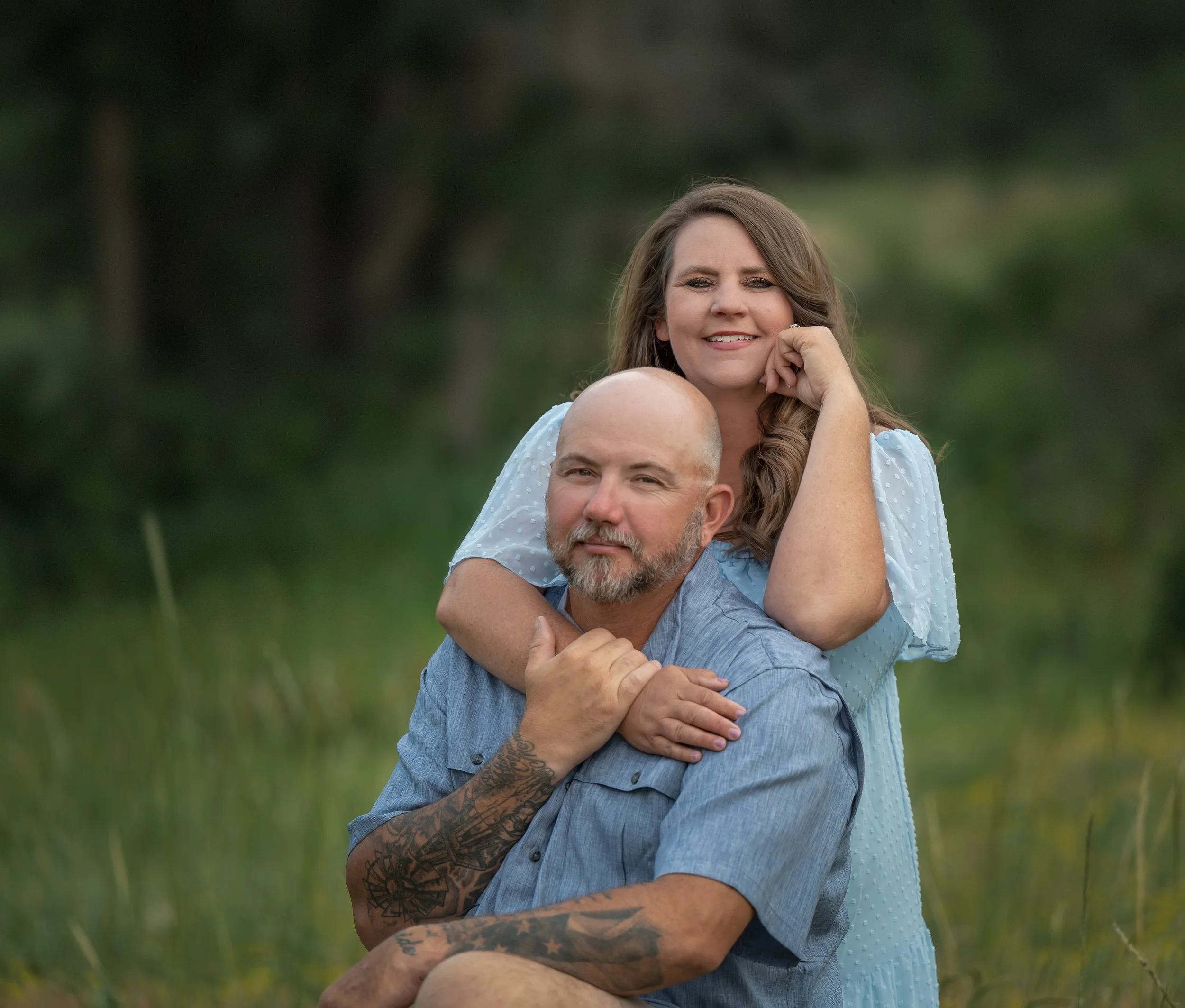 A couple outdoors in a grassy area with trees in the background, with the woman standing behind the man, resting her chin on her hand and smiling, while the man is seated, with her arms around his shoulders.