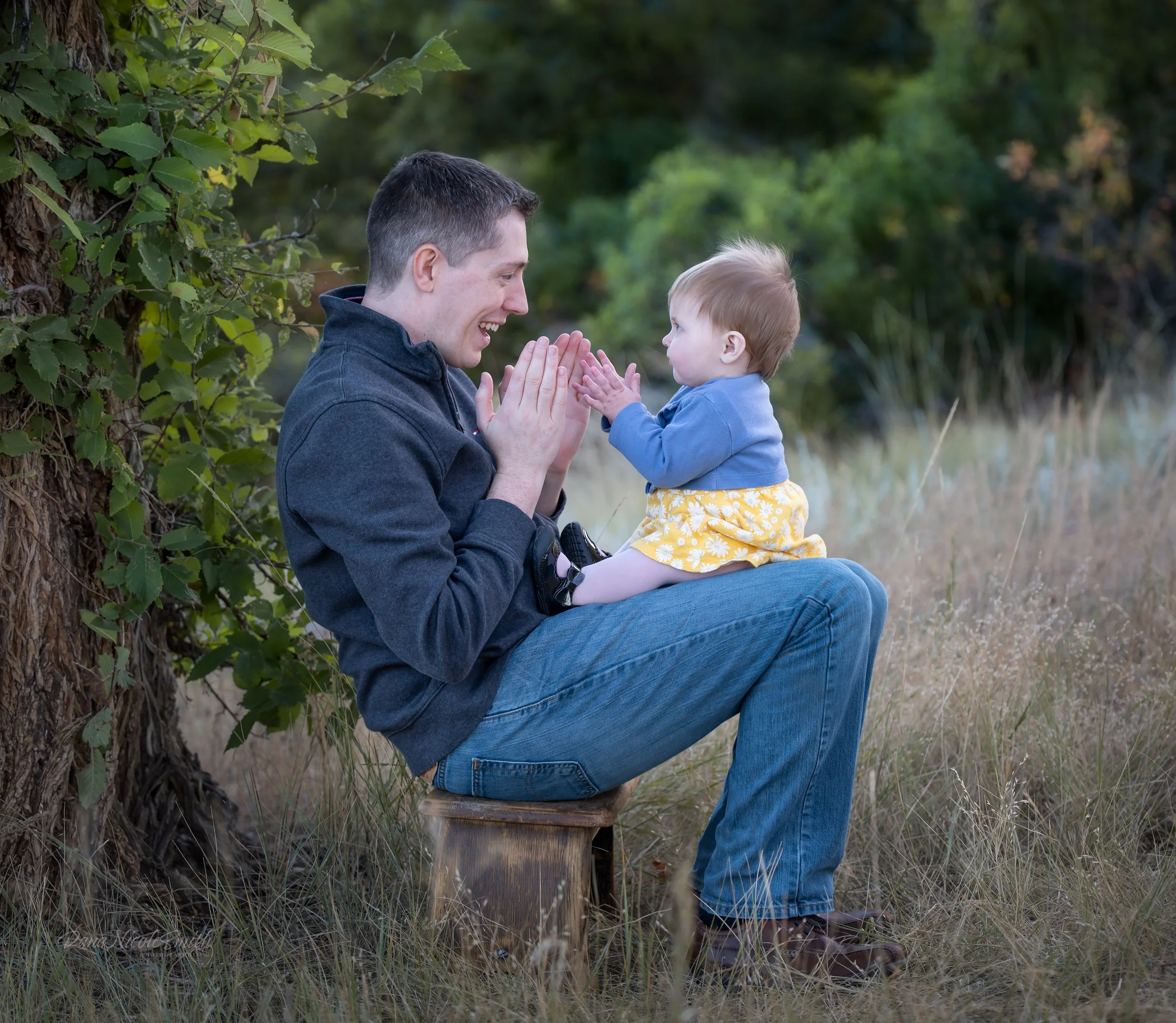 A father and young daughter sitting on a small wooden stool outdoors, playing a clapping game and smiling at each other, surrounded by grass and trees.