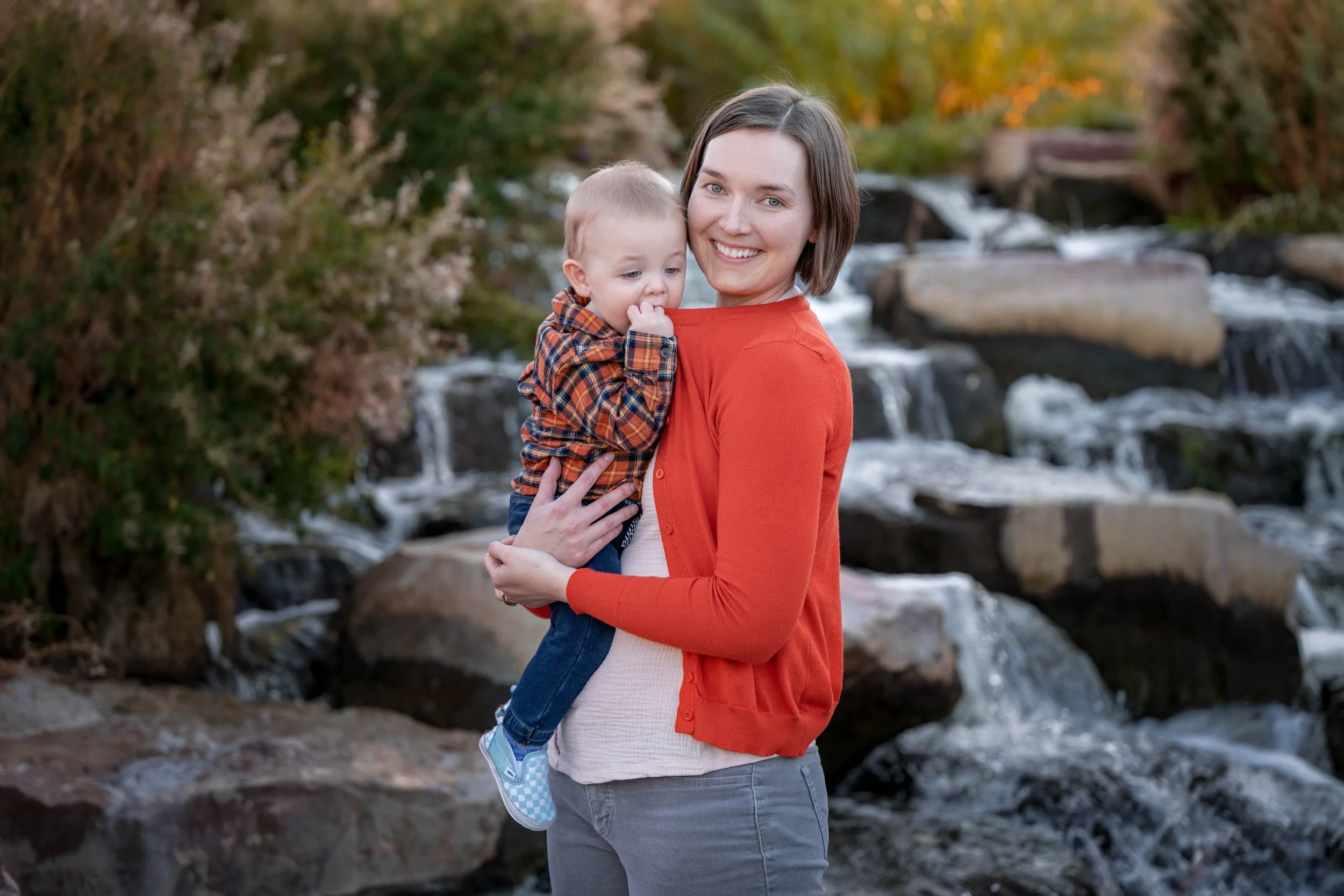 A woman holding a young boy in front of a flowing creek with rocks and autumn foliage.