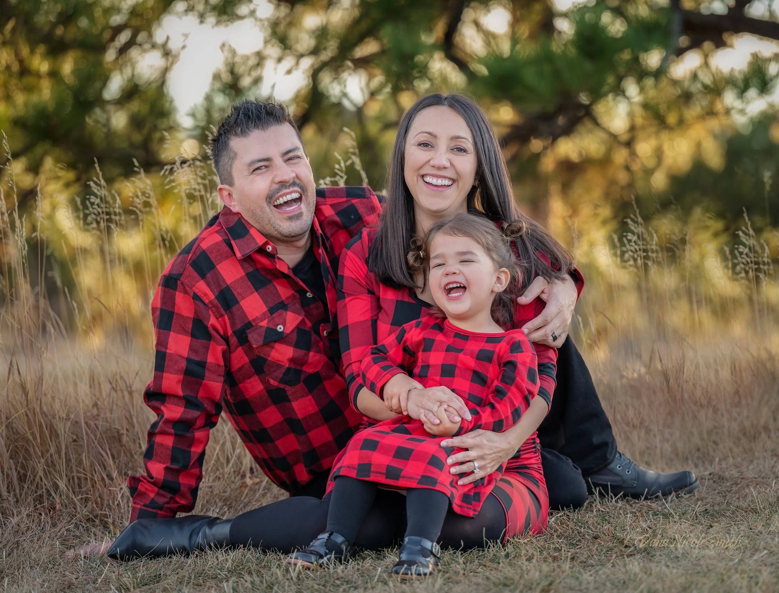 Family of three in matching red and black plaid clothing, sitting outdoors on grass with trees in the background, smiling and laughing together.