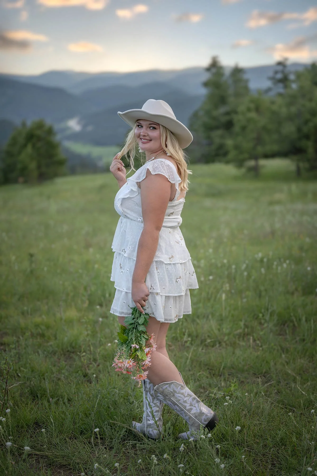 A young woman in a white, ruffled summer dress and cowboy boots, holding a bouquet of flowers, standing in a grassy field with mountains in the background during sunset.
