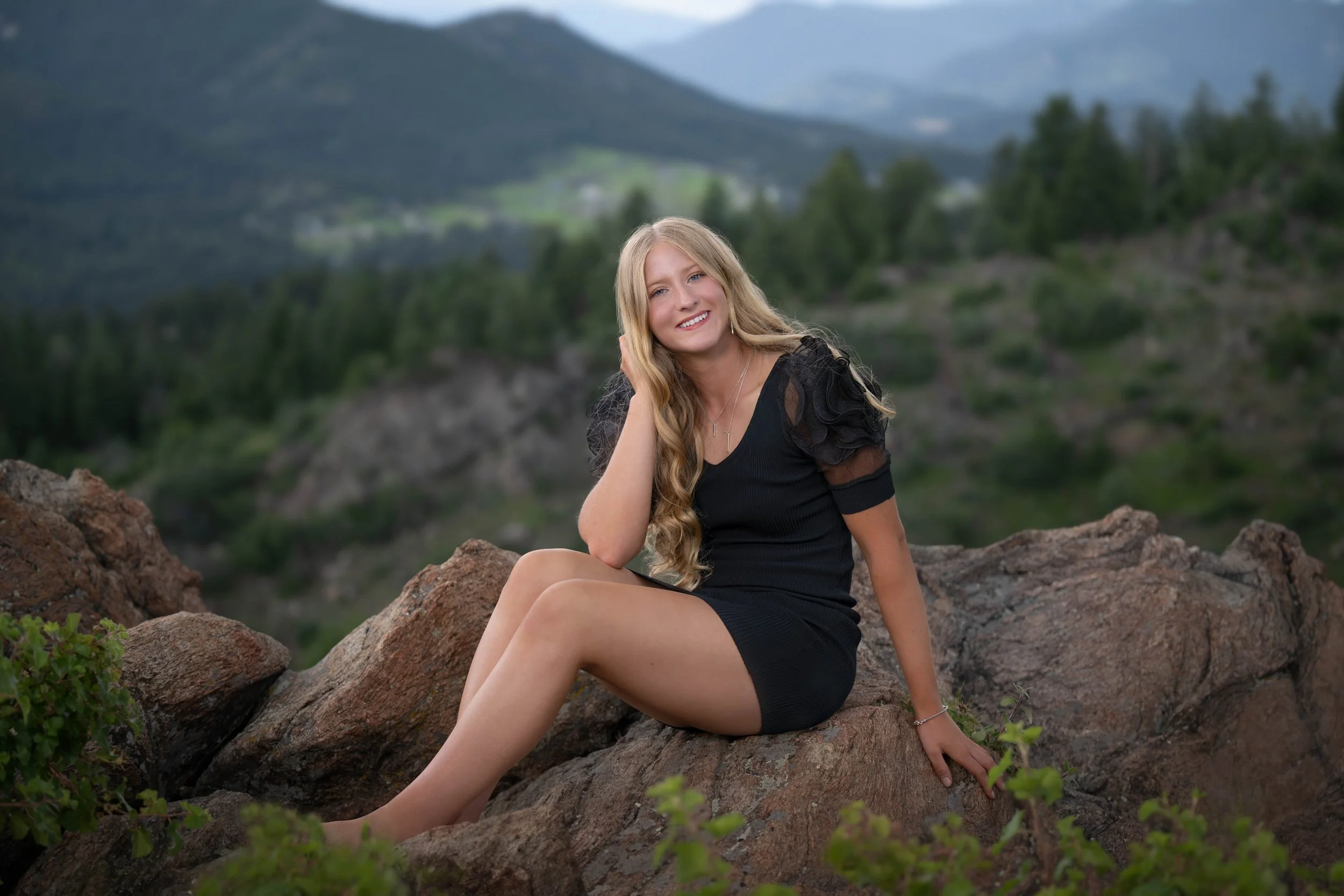 A young woman with long blonde hair, wearing a black dress with puffy sleeves, sitting on a large rock outdoors with a mountainous landscape and green trees in the background, smiling at the camera.