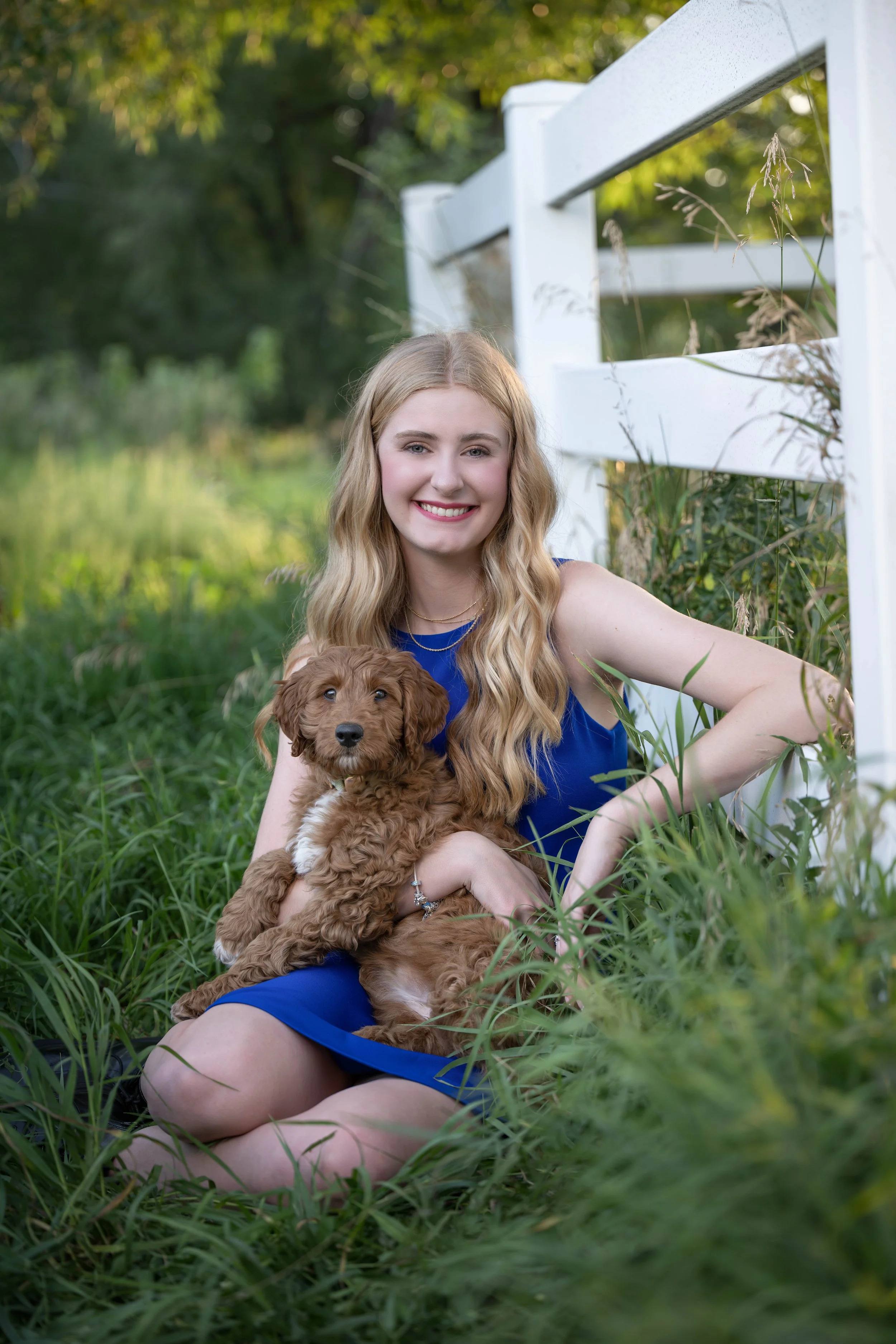 A young woman with long blonde hair wearing a blue dress sitting outdoors on grass, holding a curly-haired brown puppy in her lap, smiling at the camera, with a white fence and green trees in the background.