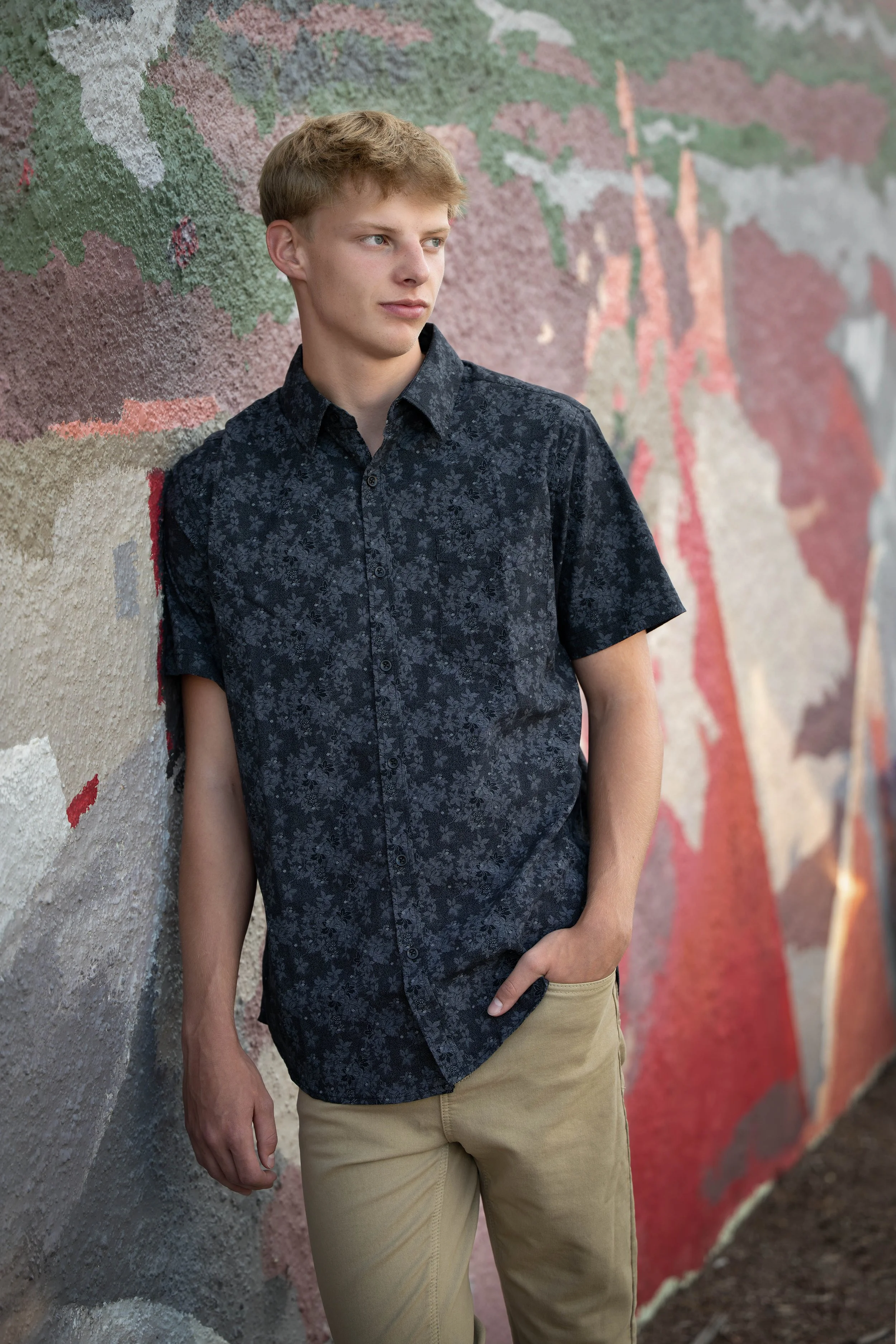 A young man with blonde hair and blue eyes standing against a colorful mural wall with a foggy, contemplative look, wearing a black patterned short-sleeve shirt and beige pants.