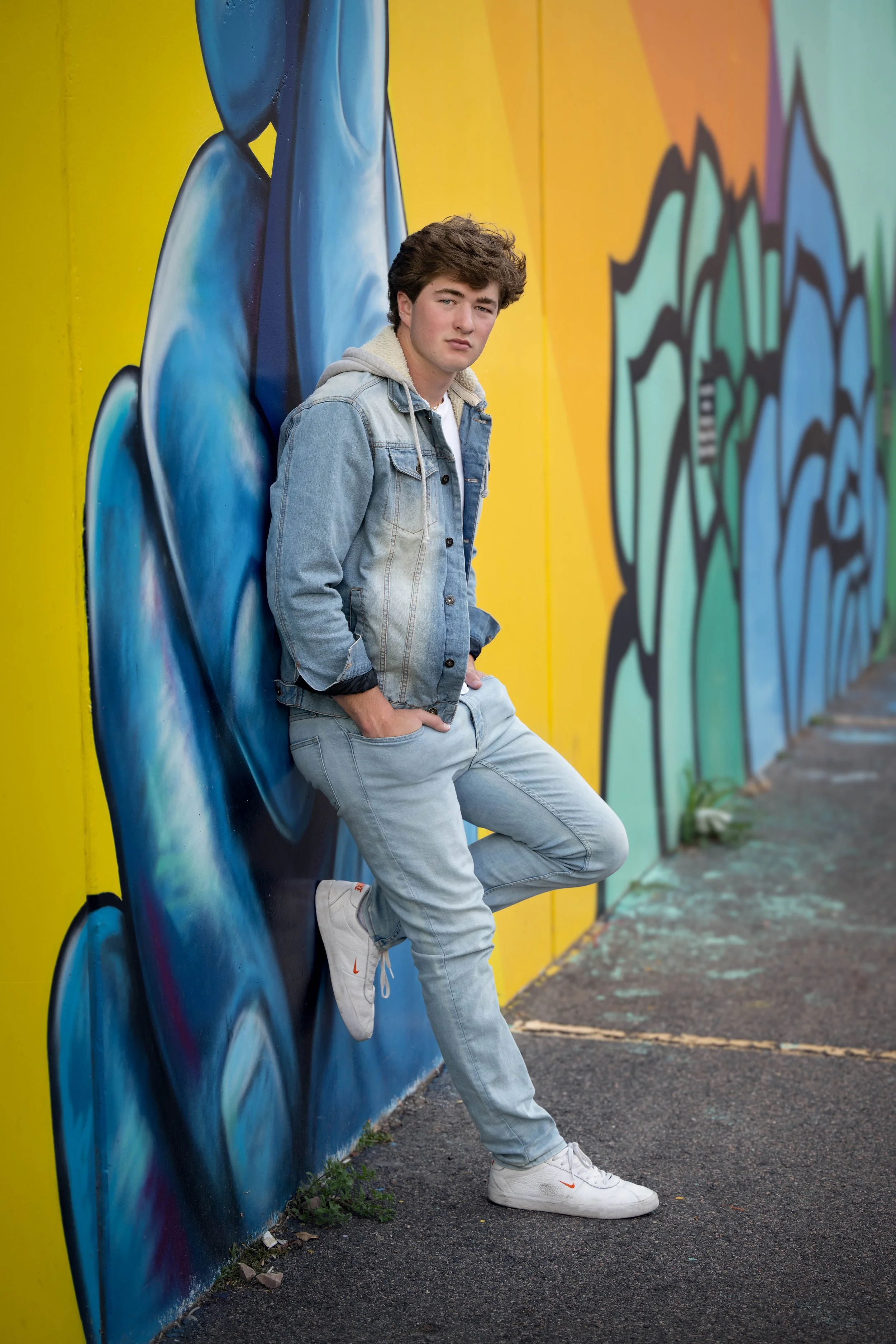 Young man in denim jacket and white sneakers leaning against a graffiti-covered yellow wall.