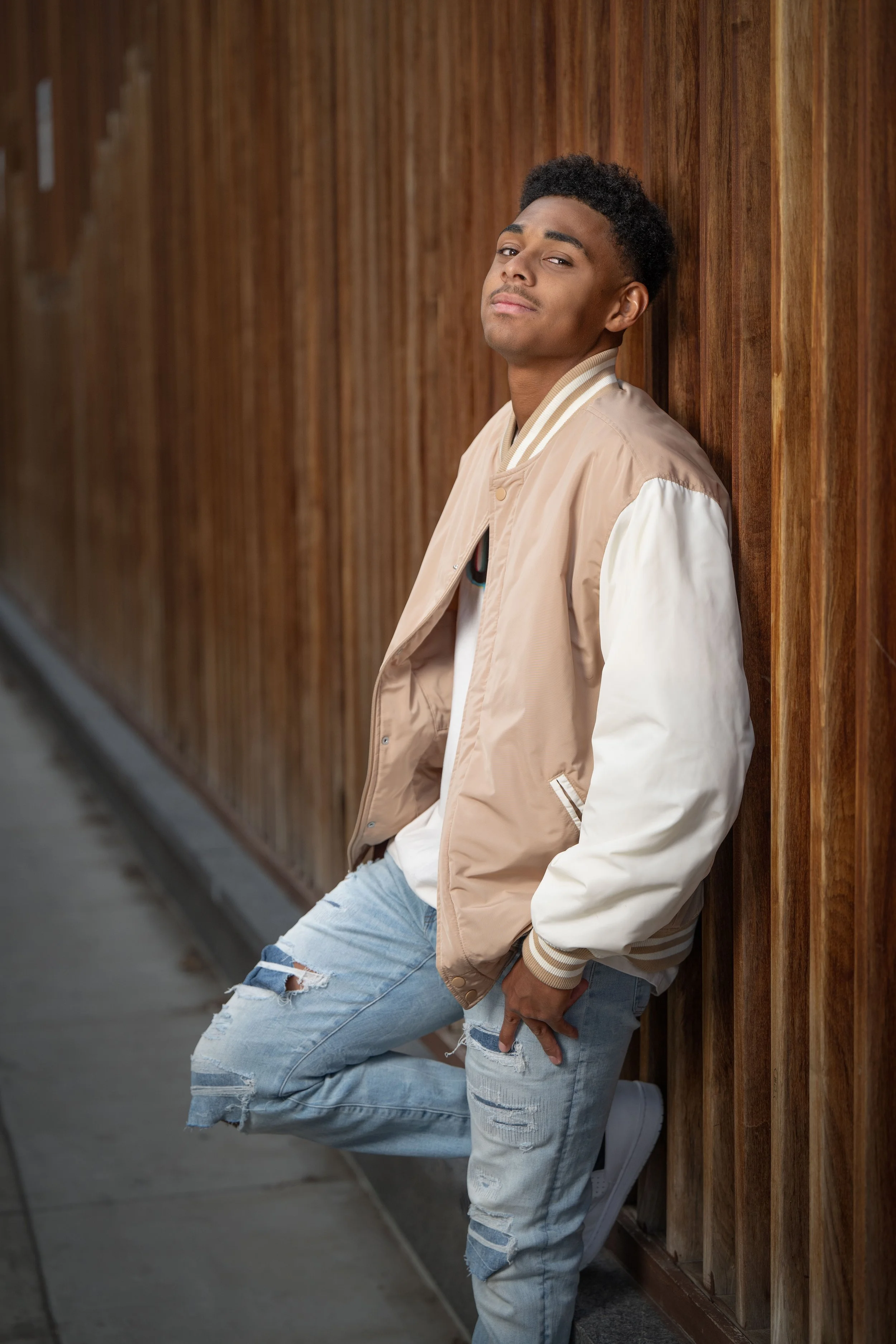 A young man leaning against a wooden wall outdoors, wearing a beige and white varsity jacket, distressed jeans, and white sneakers.