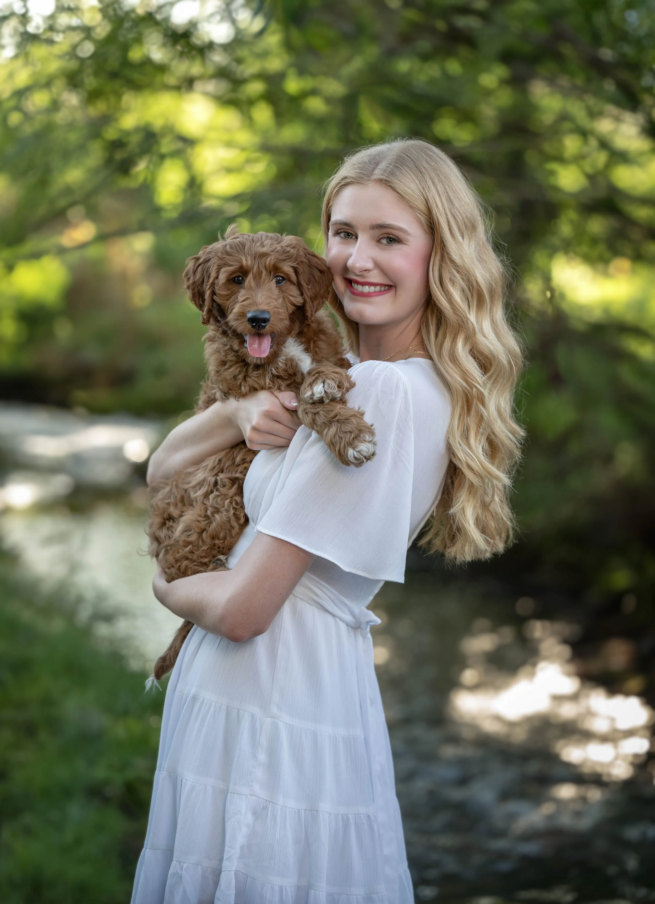 A young woman with long blonde hair smiling and holding a brown puppy in a green outdoor setting with trees and a stream.