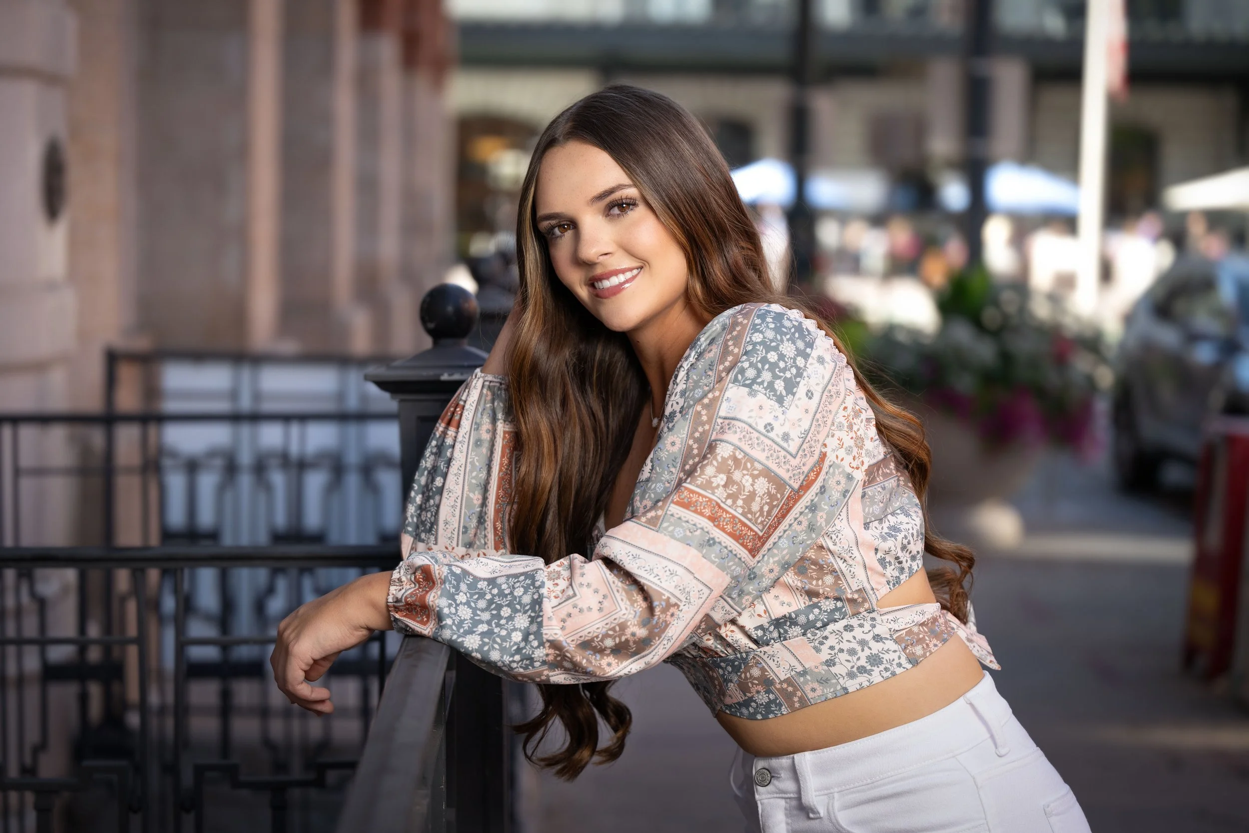 Young woman with long brown hair smiling outdoors on a city street, leaning on a black metal railing, wearing a patterned long-sleeve top and white pants.