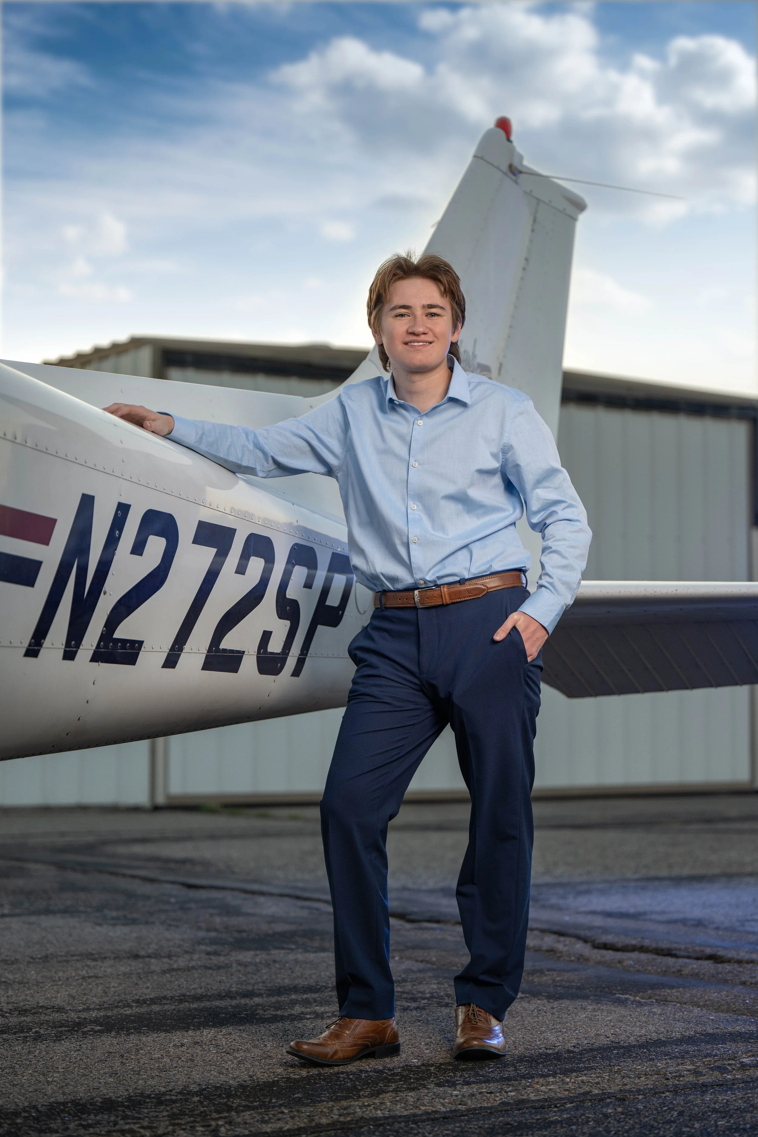 A young man in business casual attire standing next to a small white airplane, outdoors on a tarmac with hangars and a cloudy sky in the background.