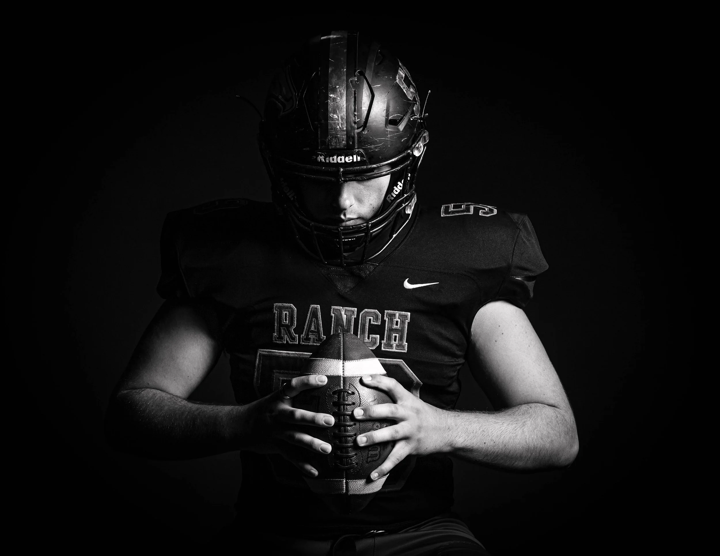 Black and white photo of a football player wearing a helmet and uniform holding a football, with a dark background.