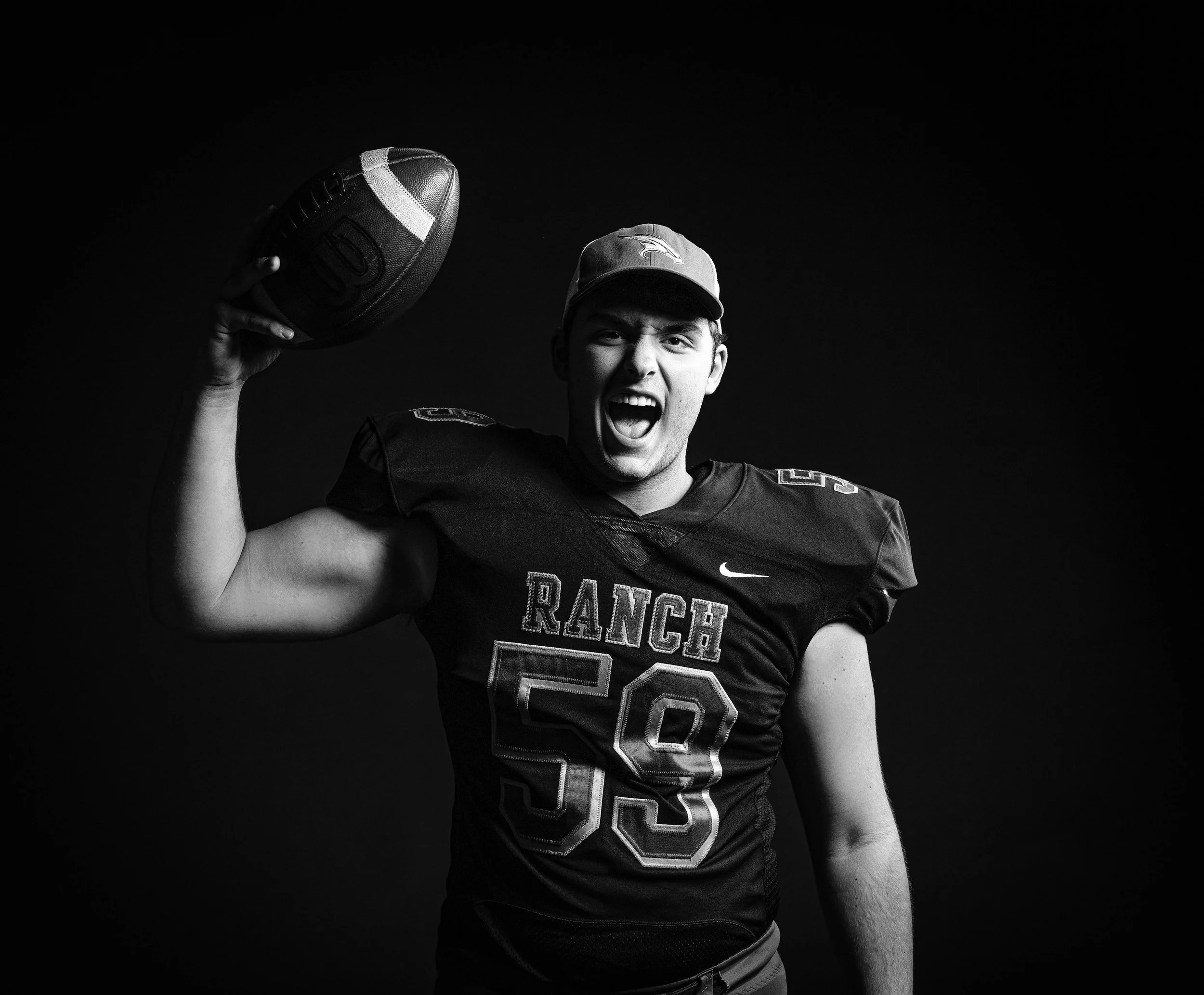 A football player wearing a jersey labeled 'RANCH 59' and a cap, holding a football in his right hand, shouting enthusiastically against a dark background.