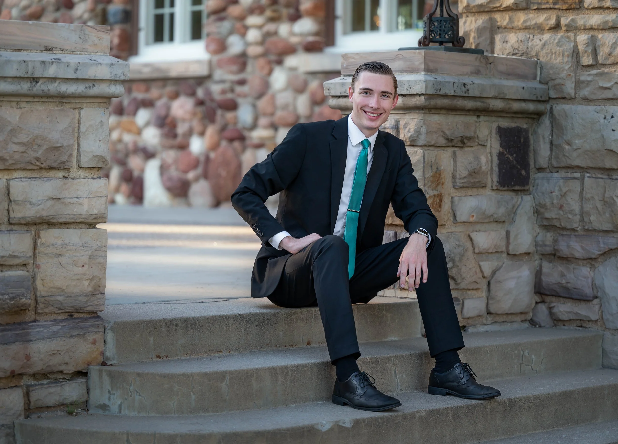 Young man in a black suit and teal tie sitting on stone steps outside a brick house, smiling at the camera.