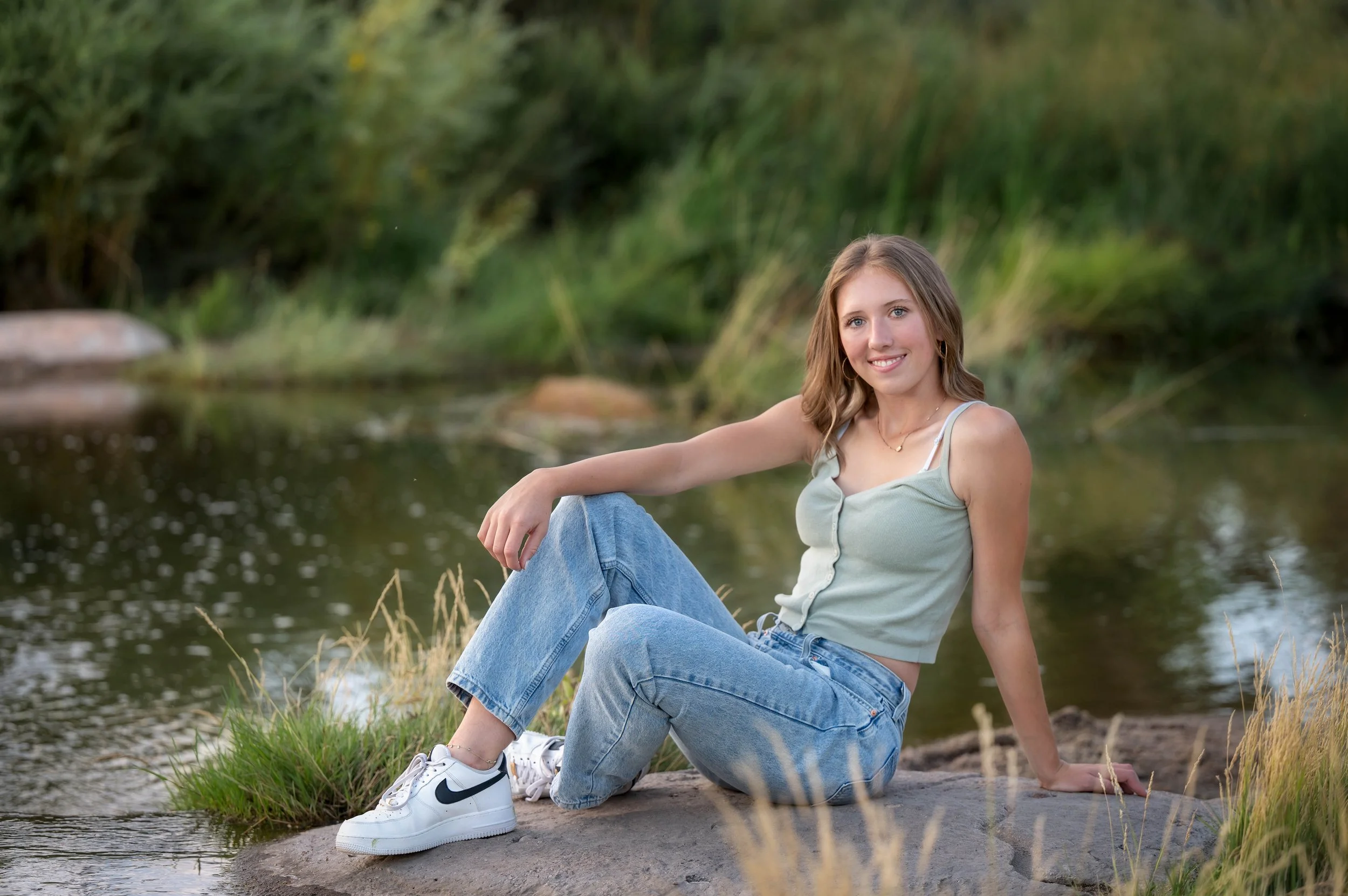 A young woman sitting on a rock by a river, wearing a light green tank top, blue jeans, and white sneakers, smiling at the camera with greenery and water in the background.