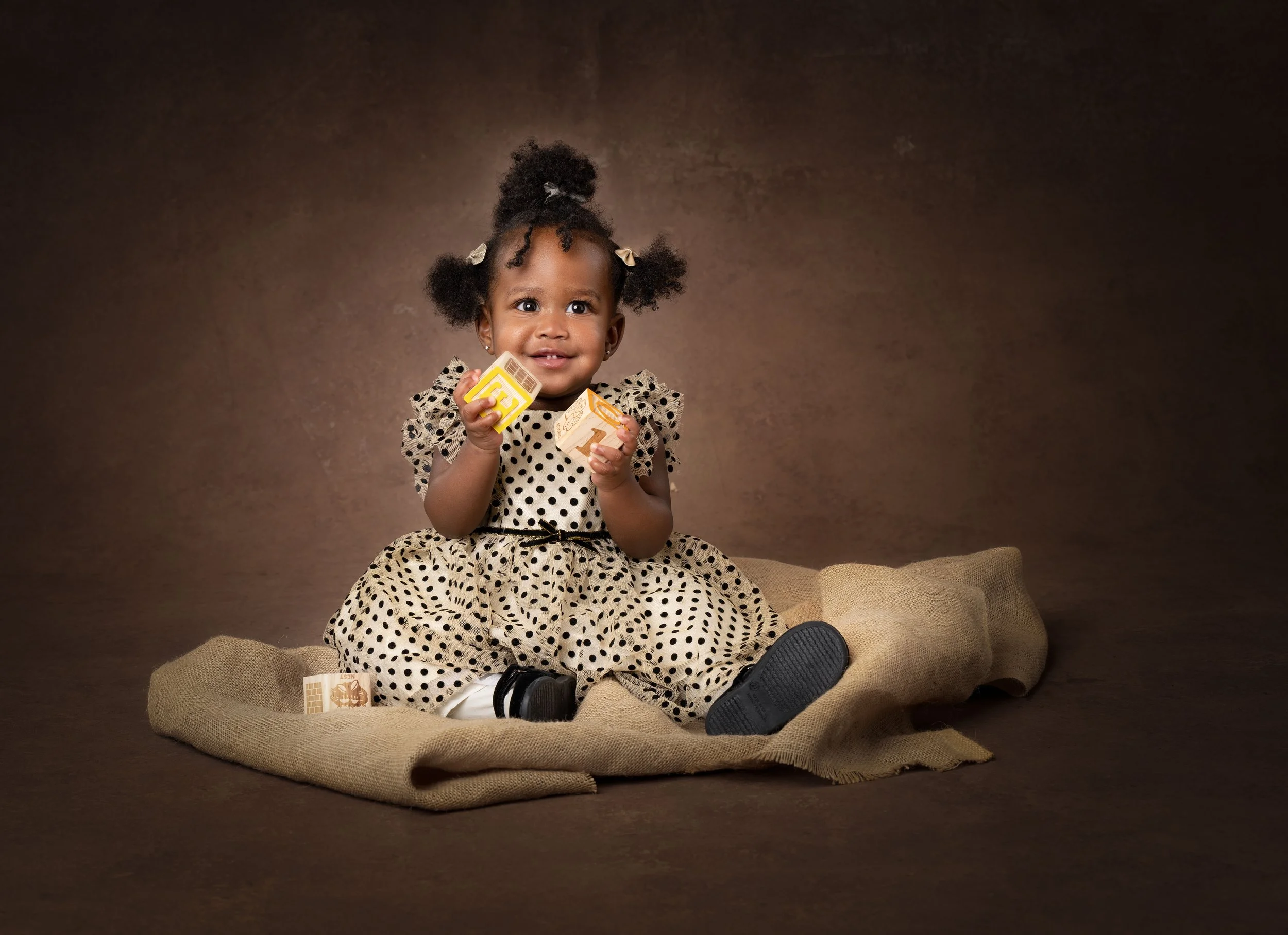 Smiling young girl with curly hair in pigtails, wearing a polka dot dress, sitting on a brown cloth, holding food containers, against a brown background.