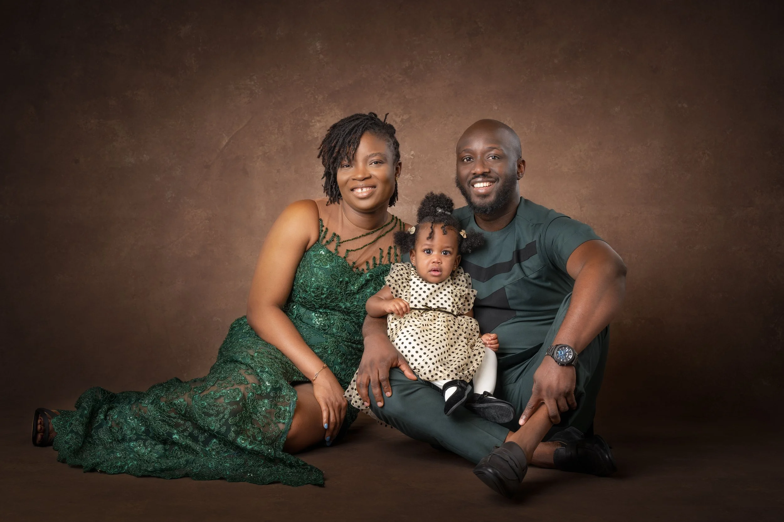 A family of three, including a woman in a green dress, a man in a dark t-shirt, and a young girl in a polka dot dress, posing together against a brown backdrop.
