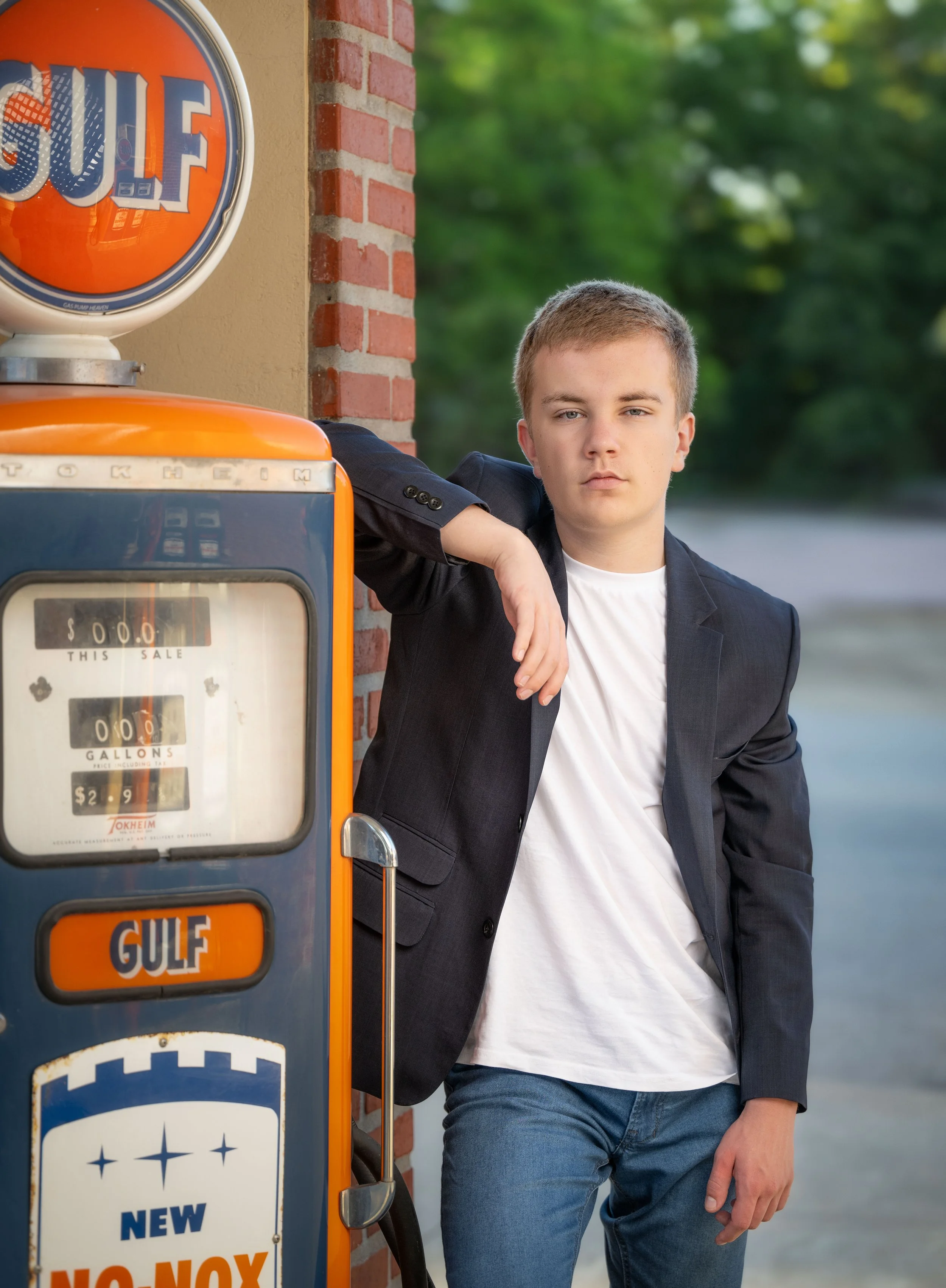 A young man in a black blazer and white T-shirt leaning against a vintage Gulf gasoline pump outside with a background of trees.