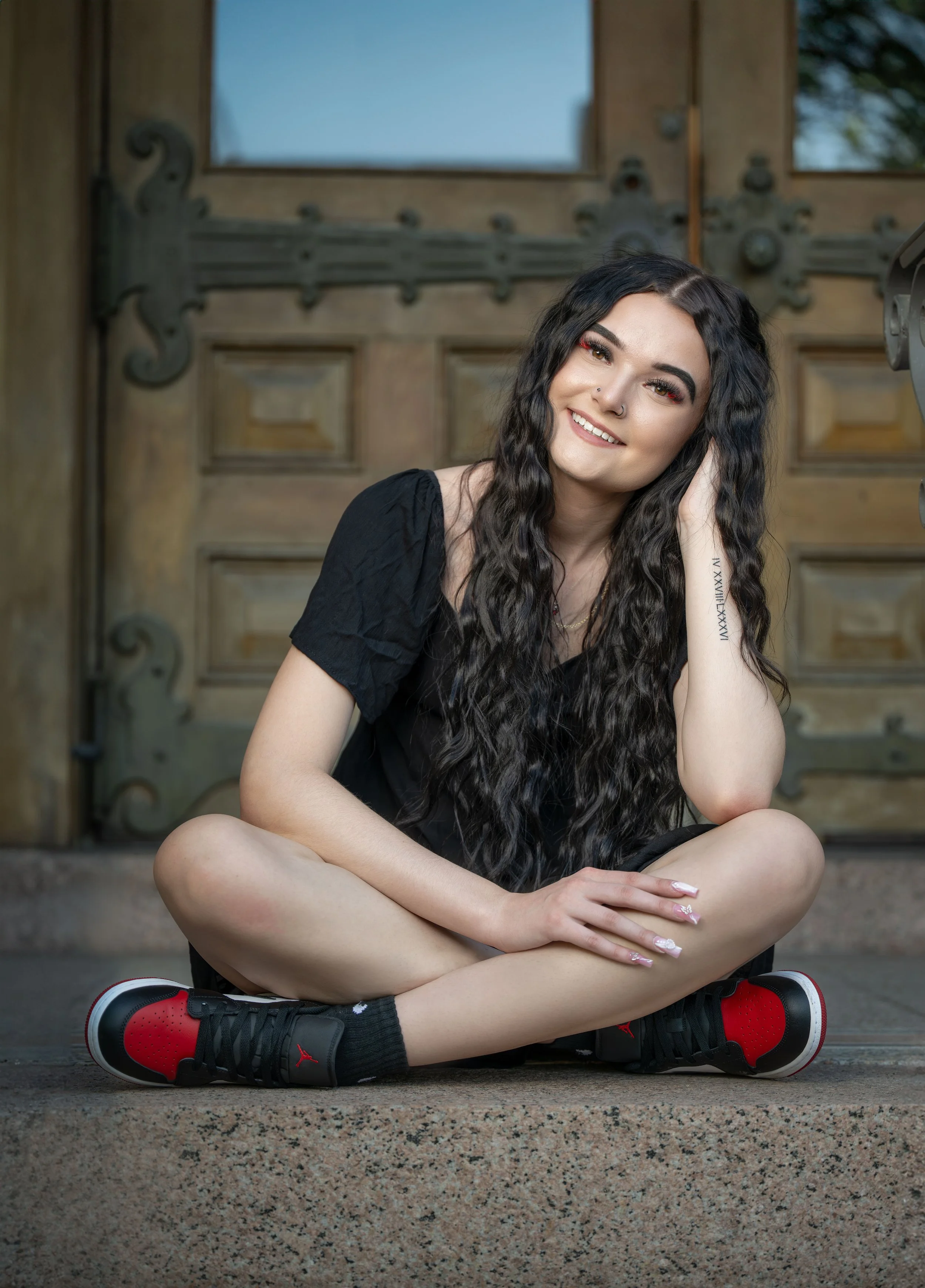 A young woman with long, curly black hair, sitting cross-legged on a concrete step in front of a wooden door. She is smiling, wearing a black t-shirt, black and red sneakers, and has a tattoo on her right forearm.