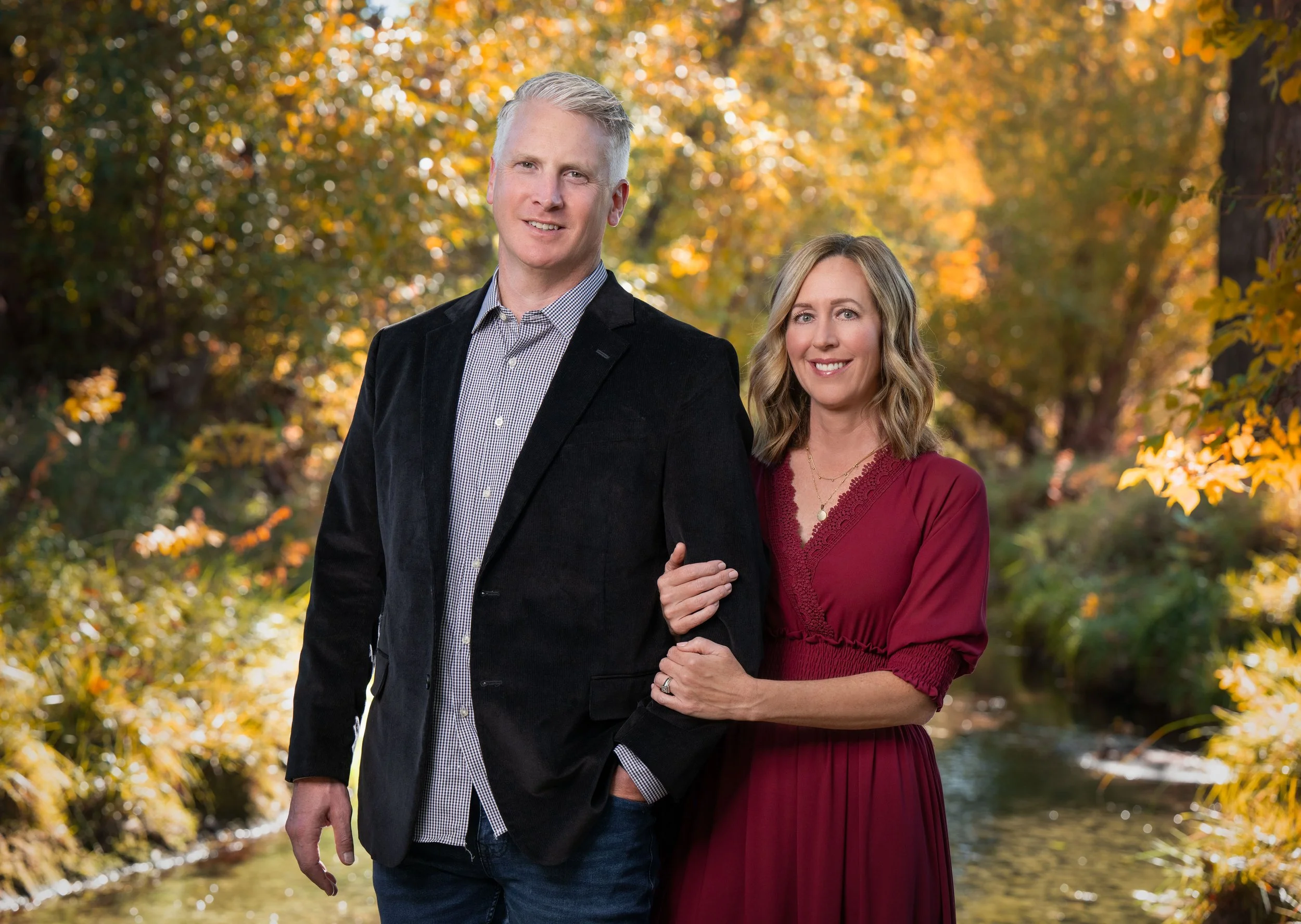 A happy couple standing outdoors in autumn, with trees and a small creek in the background. The man has short gray hair and is wearing a black blazer and a checkered shirt. The woman has shoulder-length blonde hair and is wearing a burgundy dress, ho