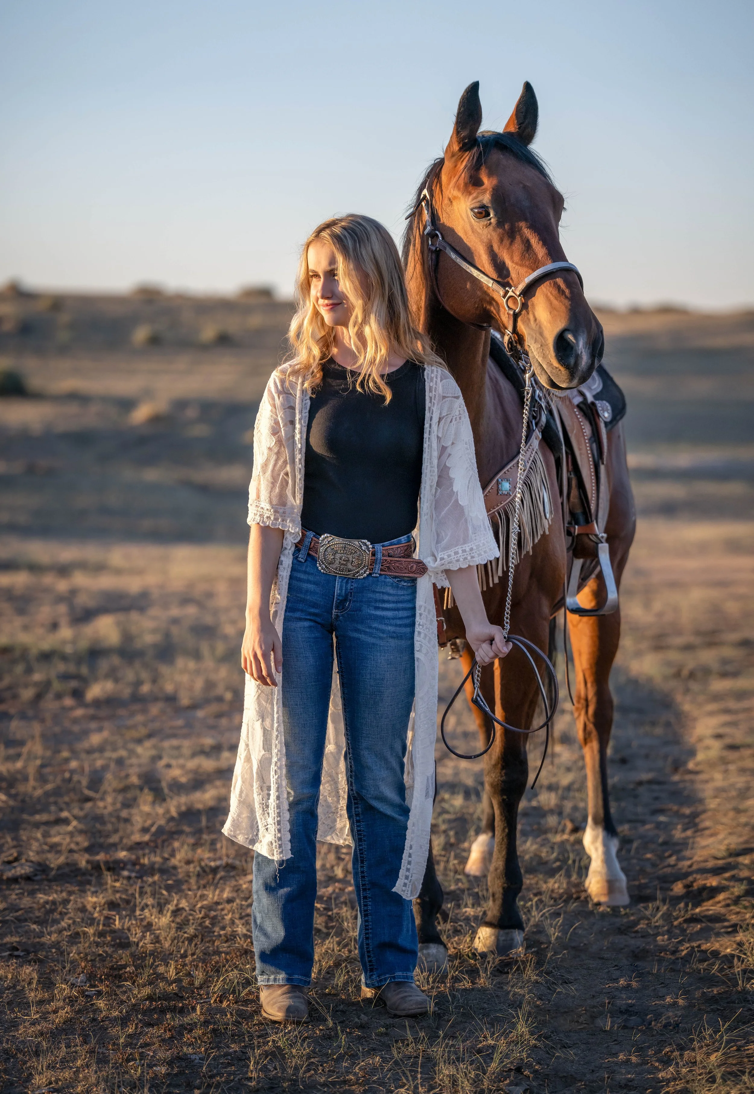 A young woman with blonde curly hair standing outdoors in a field at sunset, holding a horse by the reins. The horse is brown with a black mane, wearing a saddle, and they are both facing the right side of the image.