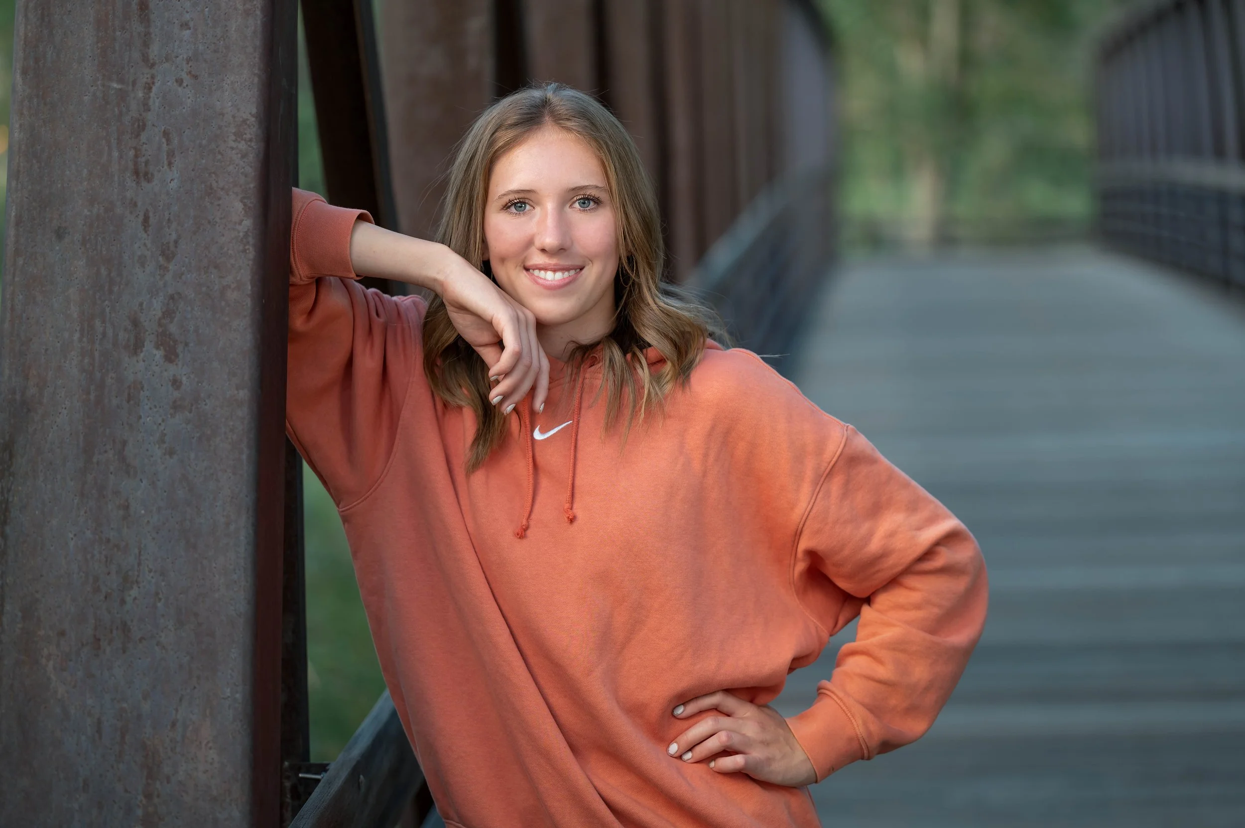 A young woman with light brown hair, wearing an orange Nike hoodie, leaning against a rusty metal structure on a wooden bridge outdoors, smiling at the camera.