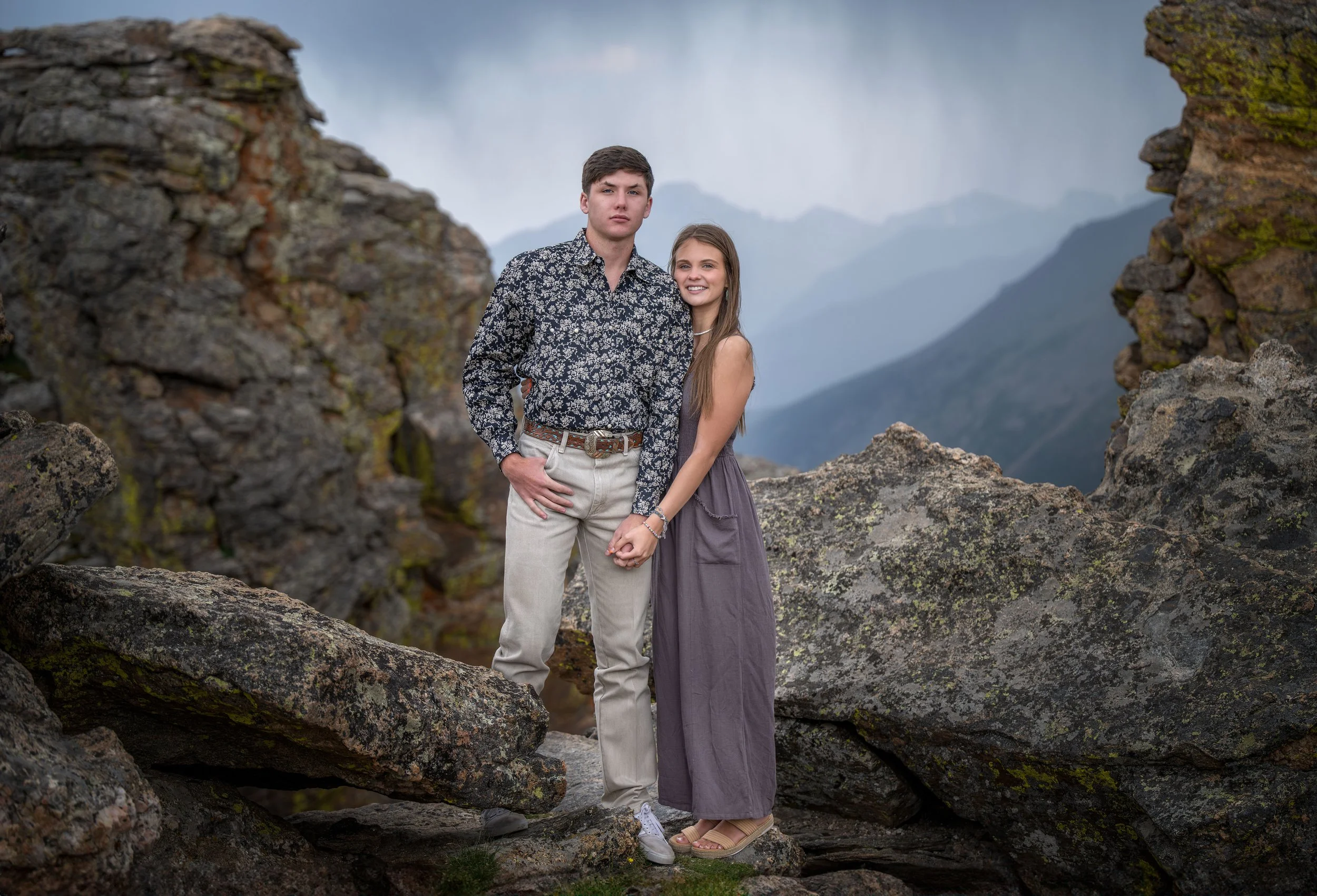 A young couple standing close together on a rocky landscape with mountains and cloudy sky in the background, holding hands and smiling.
