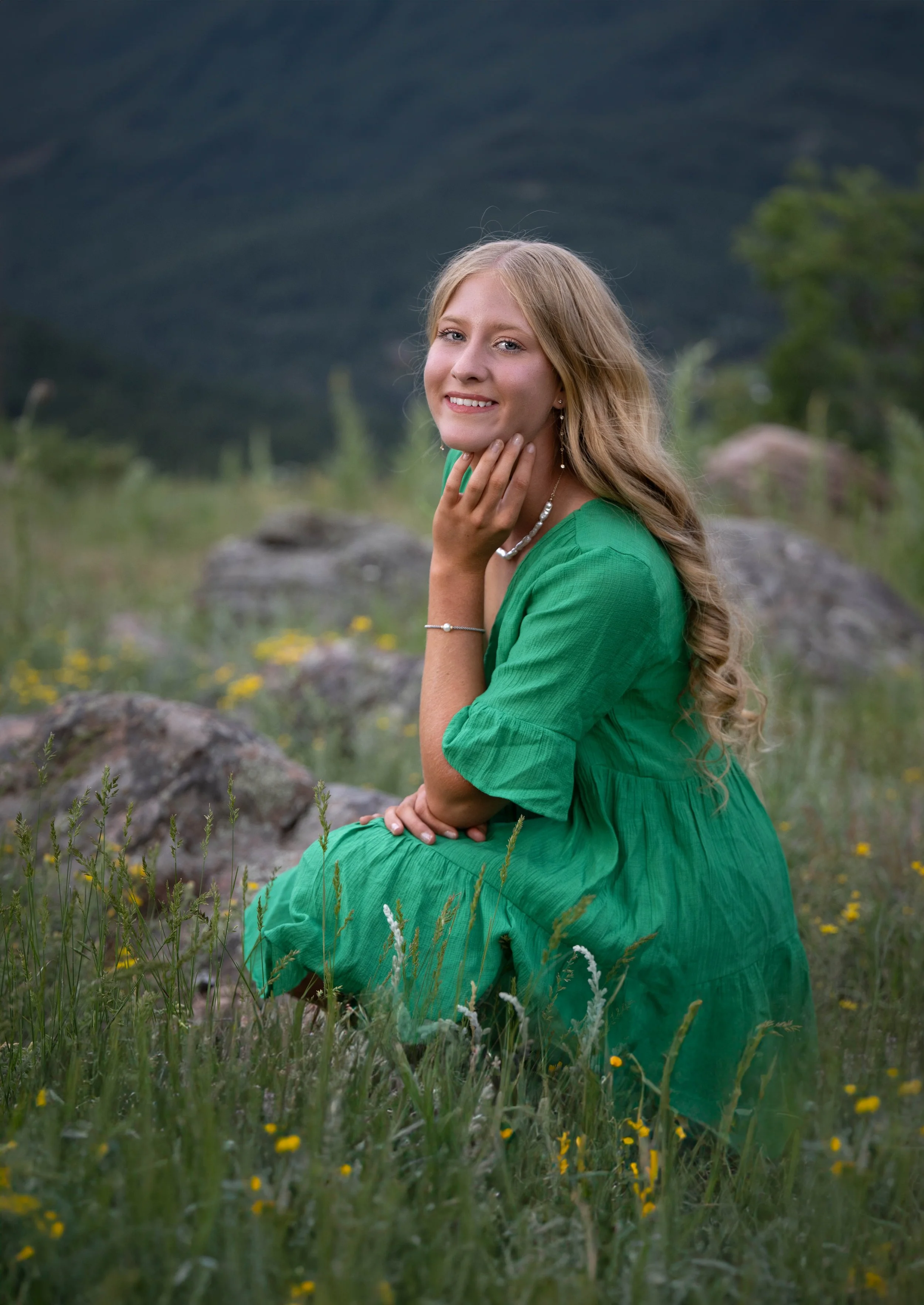 A young woman with long blonde curly hair wearing a green dress, sitting on the grass in a field with yellow flowers, with mountains in the background, smiling and looking at the camera.