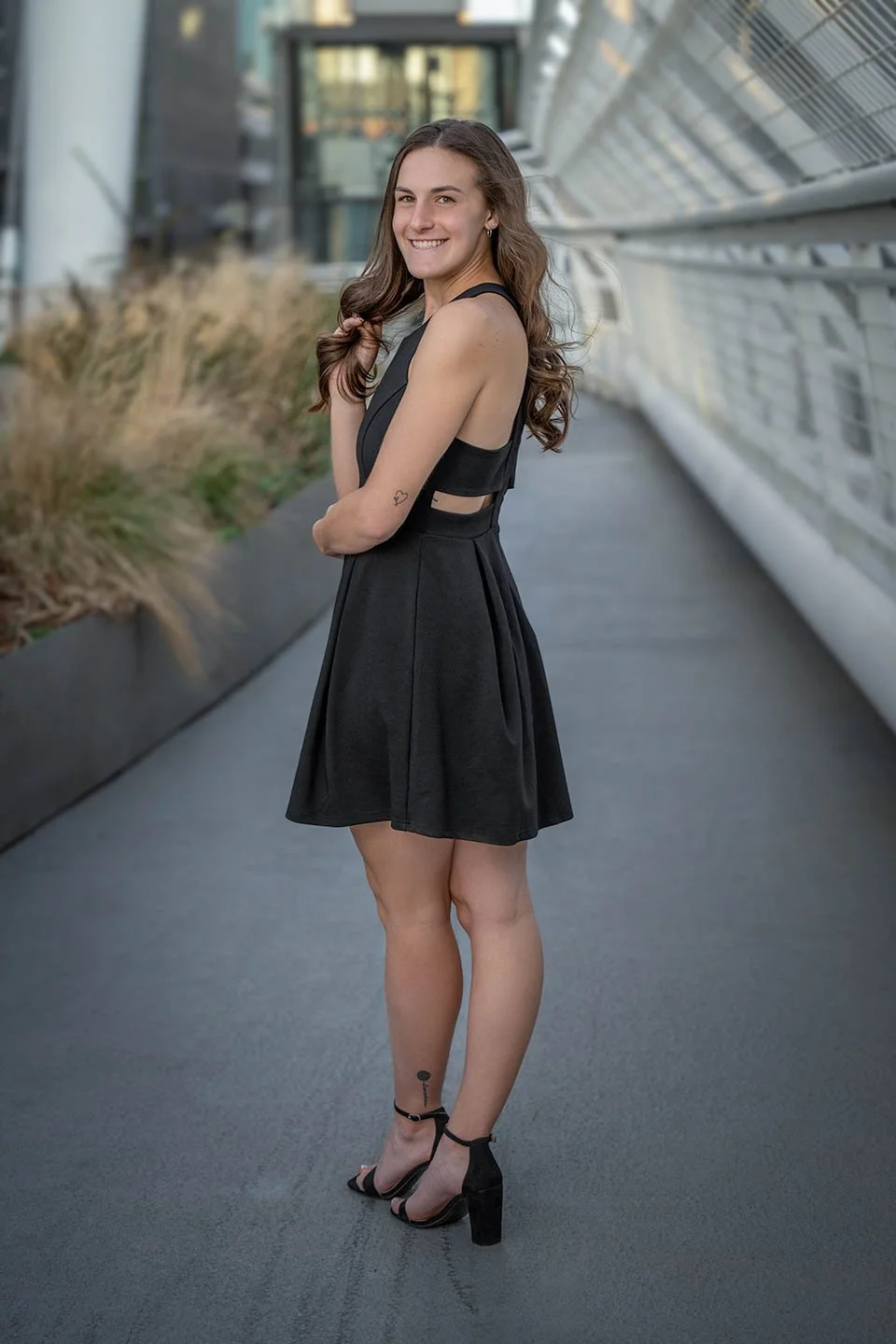 Smiling woman in a black dress and high heels standing on a city walkway.