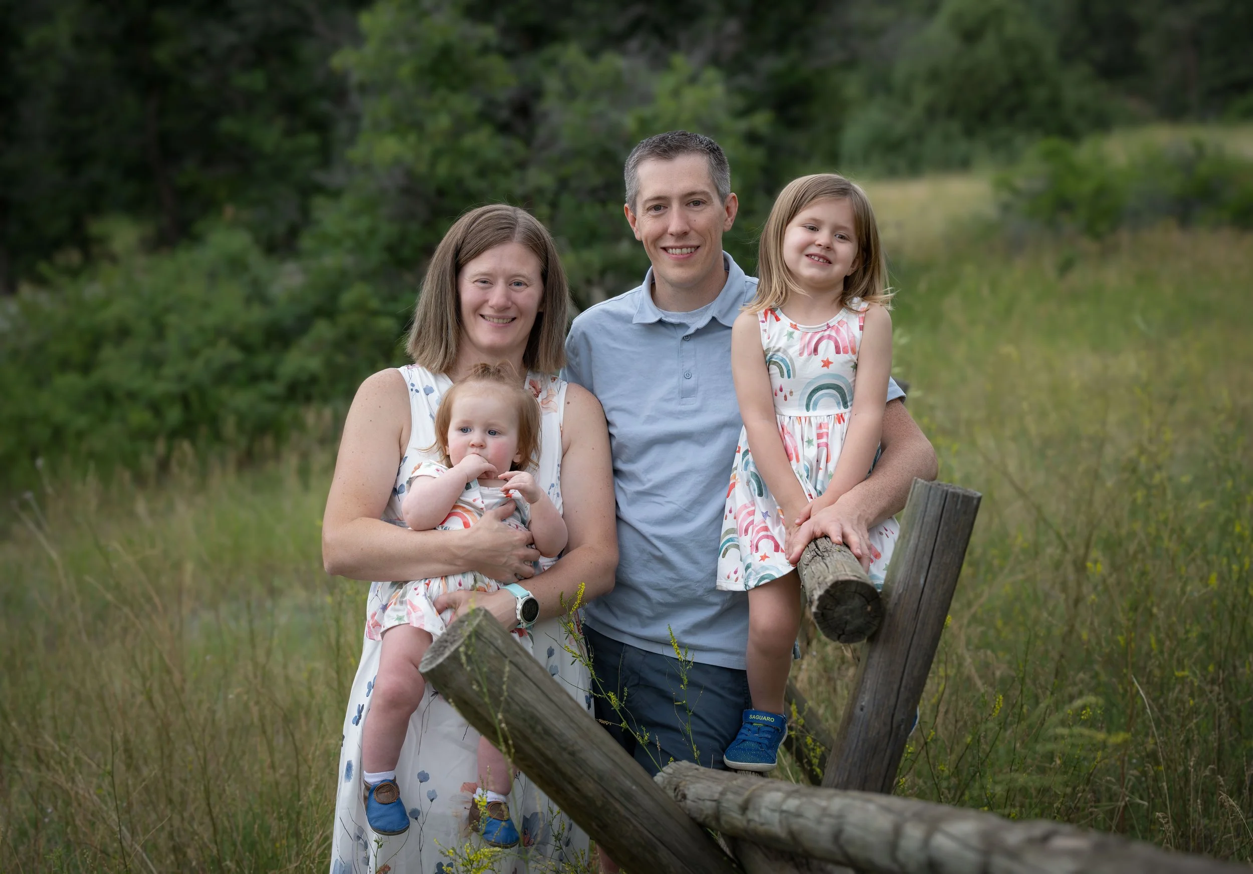 A family of four standing outdoors in a grassy field with trees in the background. The mother is holding a toddler girl, and a young girl is sitting on a wooden fence next to the father.