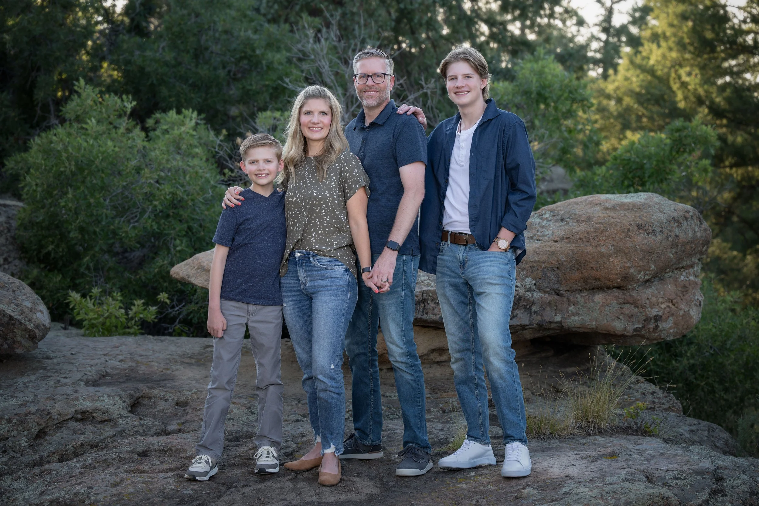 Family of five standing outdoors on rocky terrain with greenery and large rocks in the background, smiling at the camera.