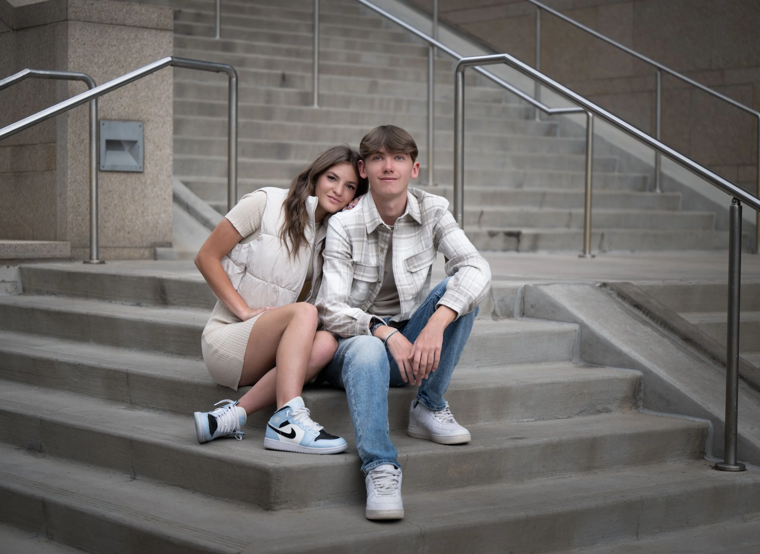 A young couple sitting on concrete stairs with metal railings in an urban setting, the girl leaning her head on the boy's shoulder.
