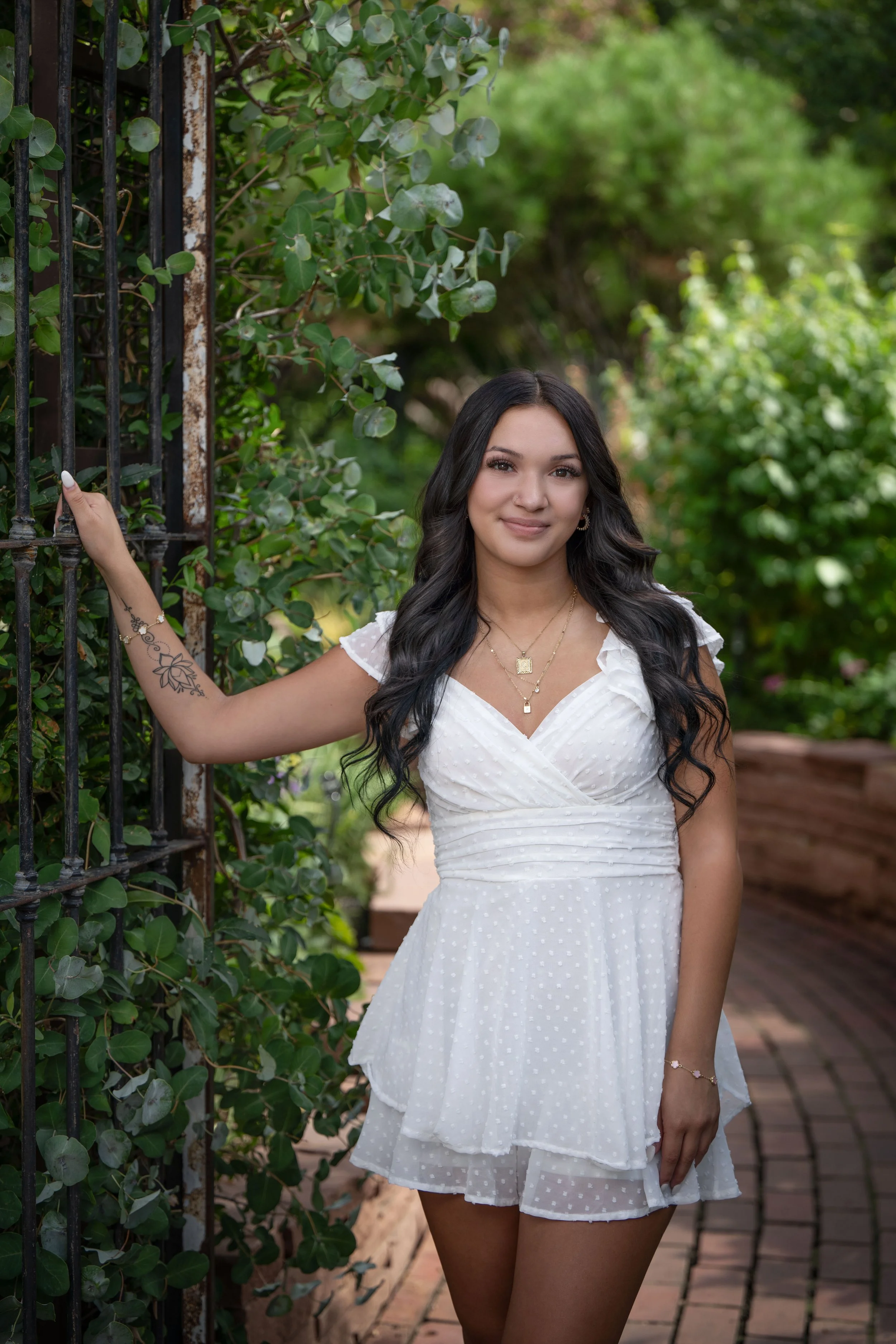 A young woman with long dark hair in a white dress standing outdoors on a brick path, holding onto a black iron gate, surrounded by greenery.