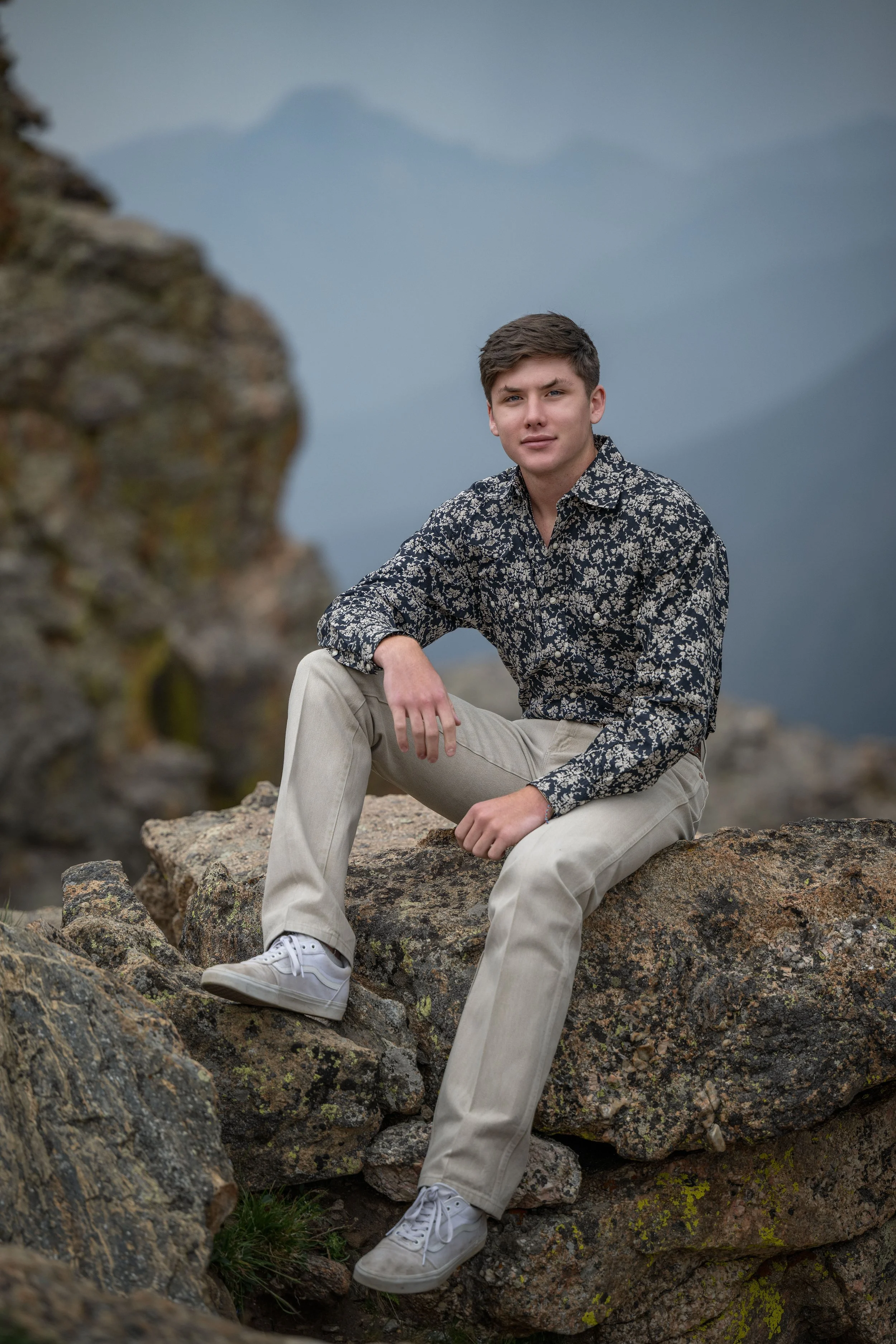 Young man sitting on rocks in a mountainous outdoor setting, wearing a patterned shirt and light-colored pants, with mountains in the background.