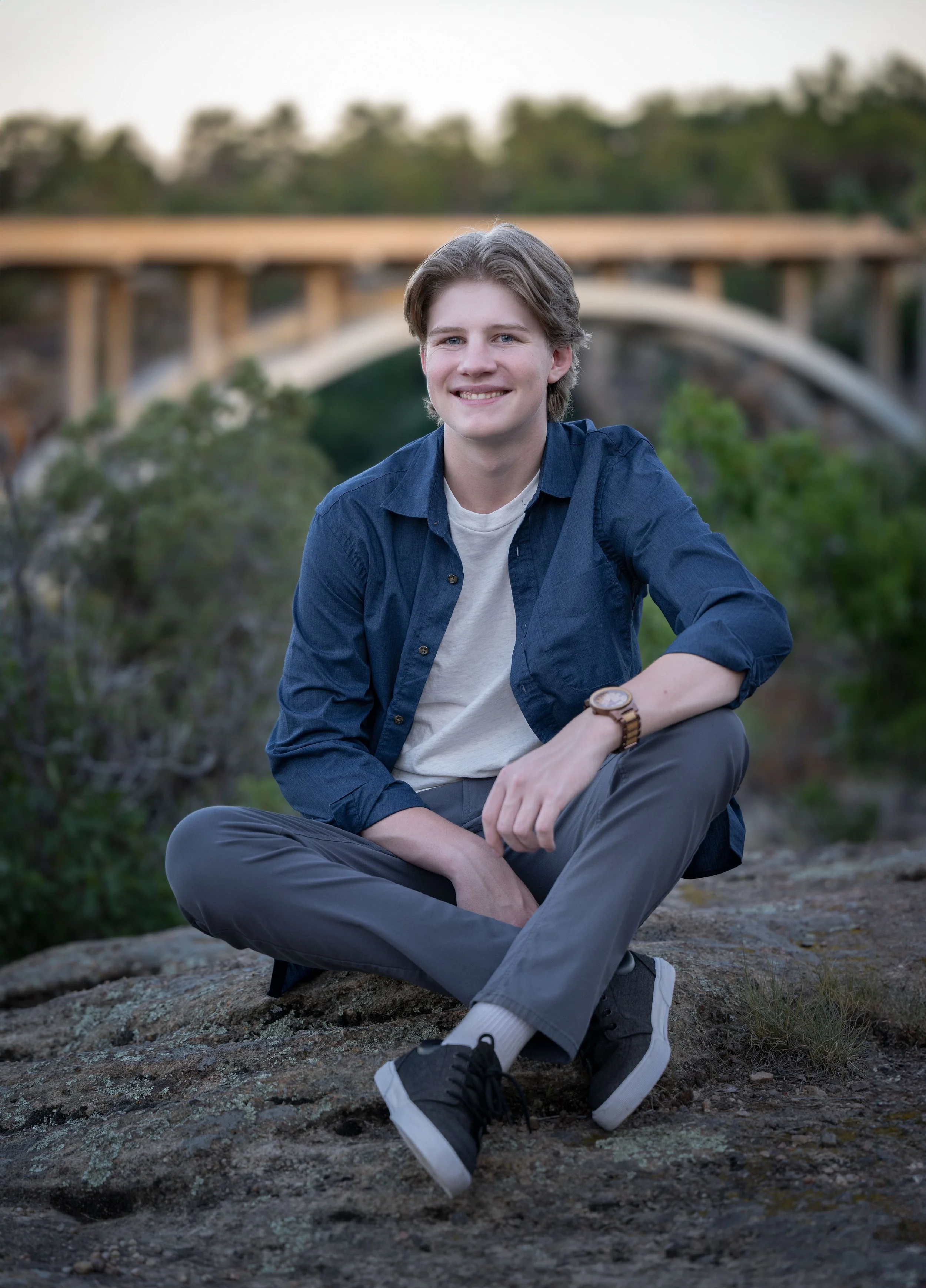 A young man with light brown hair smiling, sitting cross-legged on a rock outdoors, with a bridge and trees in the background.