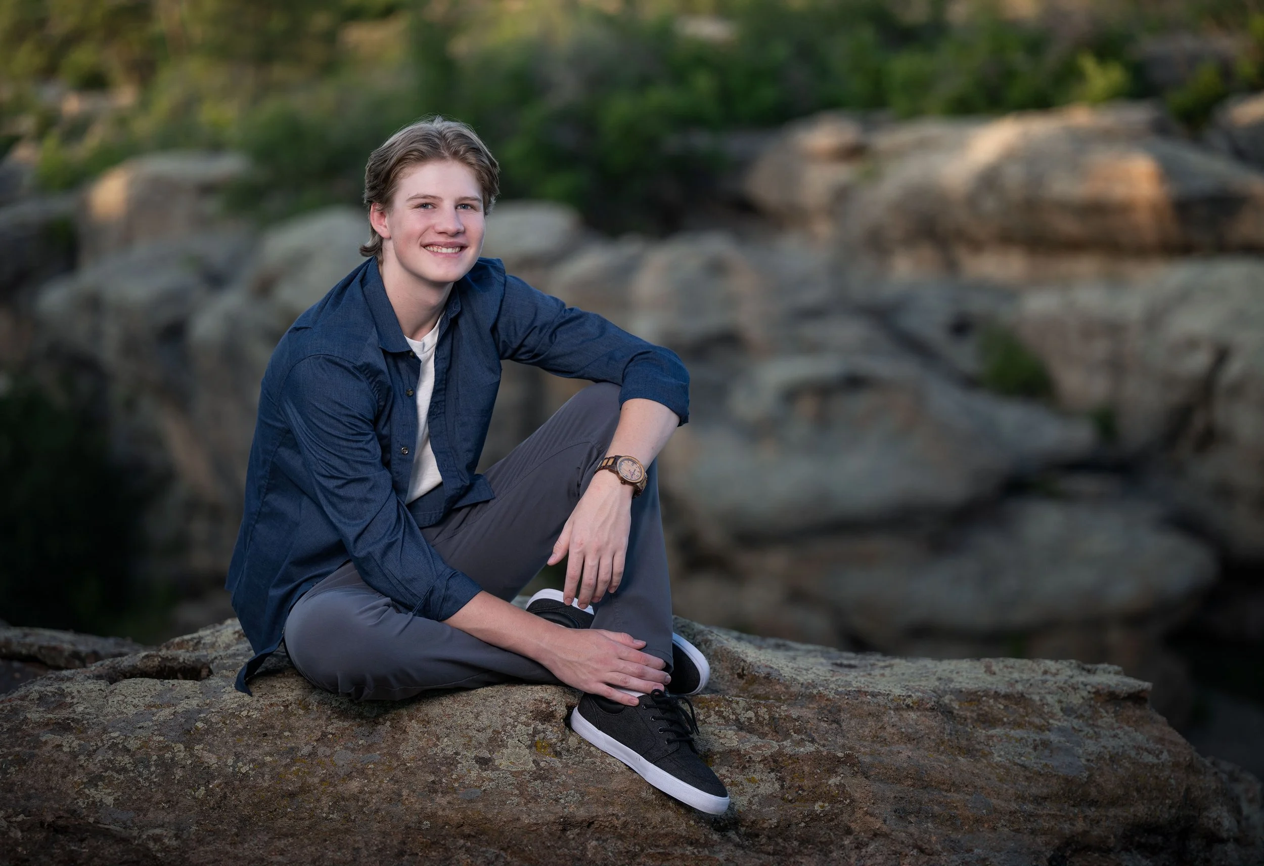 A young man with light brown hair, wearing a blue shirt, gray pants, and black sneakers, sitting on a rock outdoors with a natural rocky landscape and greenery in the background, smiling at the camera.