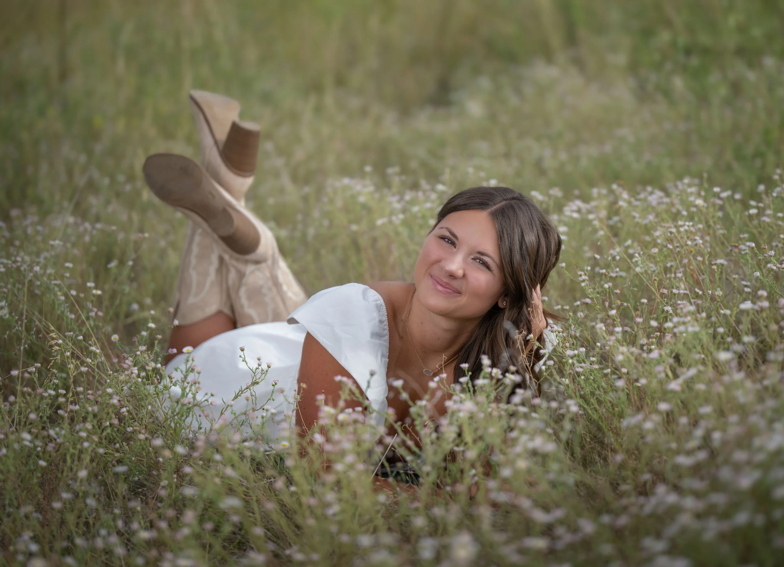 A young woman lying on her stomach in a field of small white and purple flowers, smiling and resting her head on her hand, wearing a white dress and beige patterned boots.