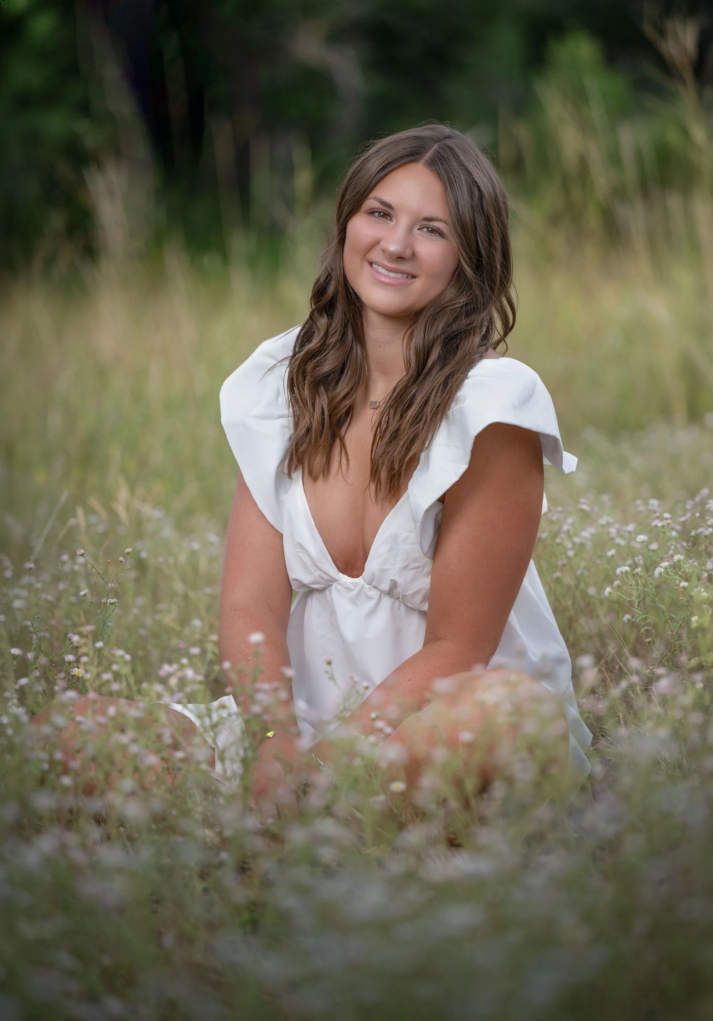 A young woman with long wavy brown hair sitting in a field of small wildflowers, wearing a white dress, and smiling at the camera.