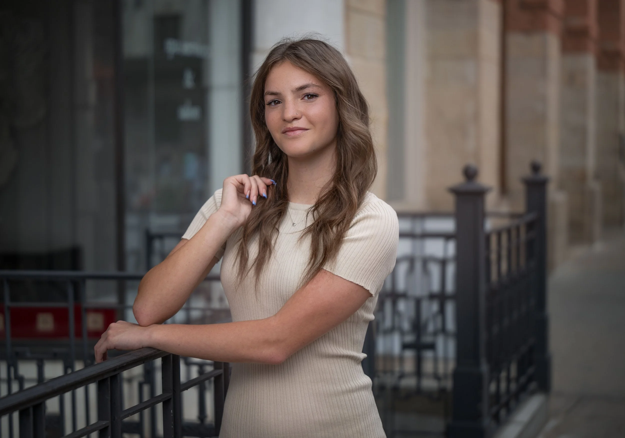 A young woman with long wavy hair and a light-colored, short-sleeved shirt standing on a city sidewalk, leaning on a black metal railing, with brick and glass buildings in the background.