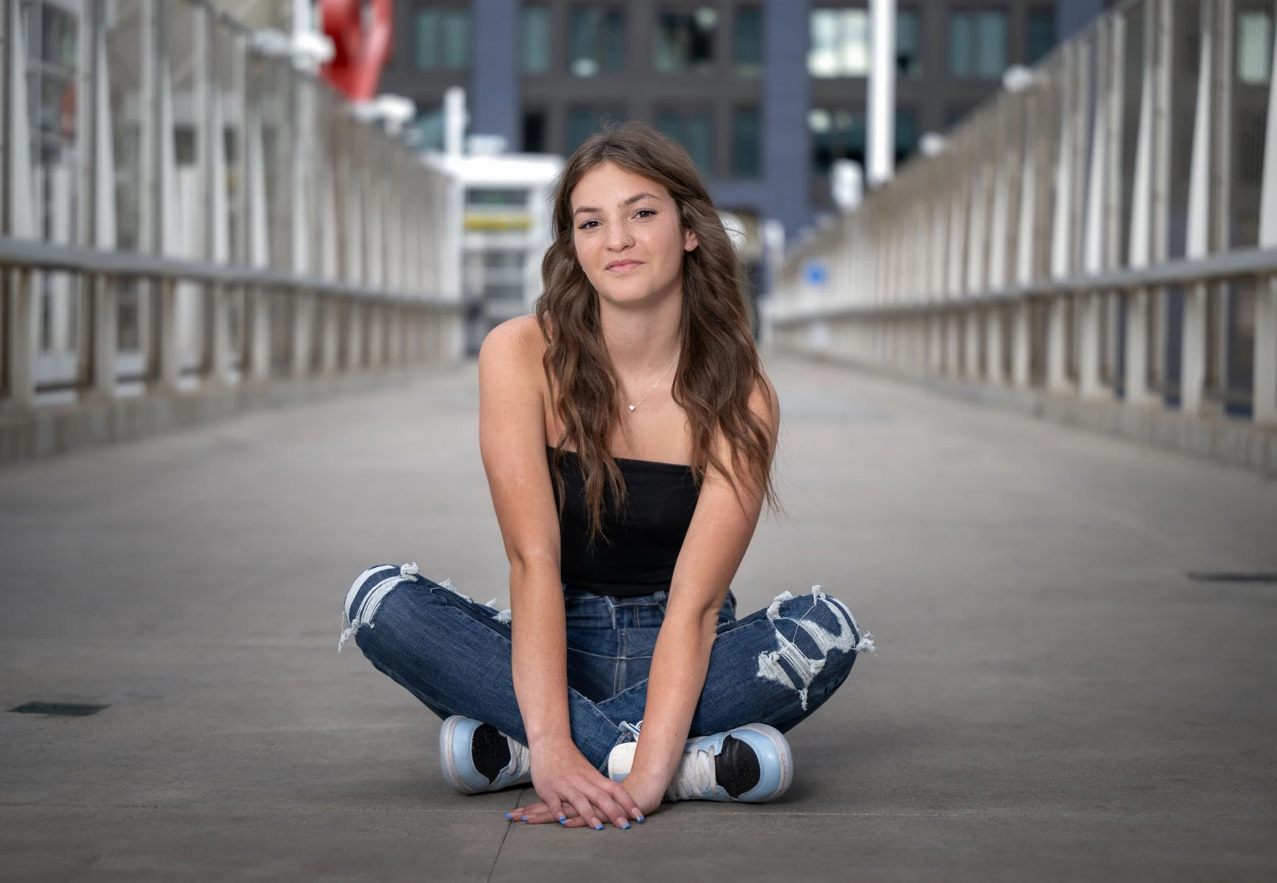 Young woman with long wavy hair sitting on the pavement in casual clothes, in an urban setting with modern buildings and railings in the background.