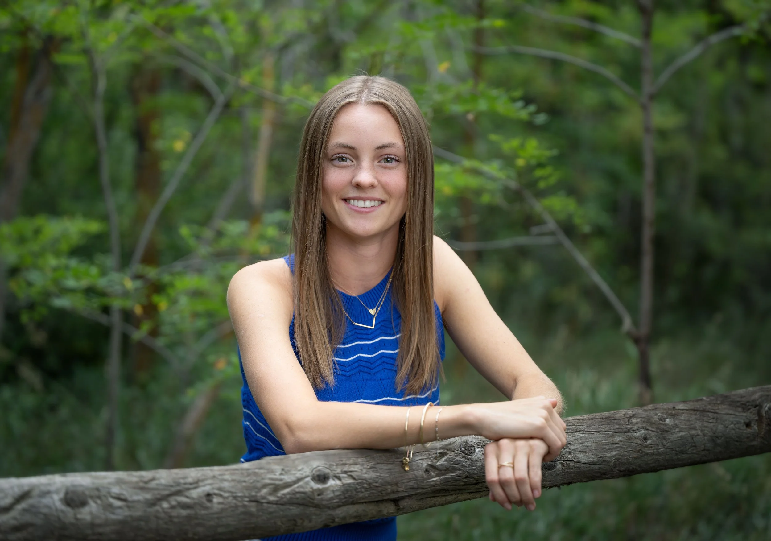 A smiling young woman with long brown hair wearing a blue sleeveless top, standing outdoors in a green forest, leaning on a wooden log.