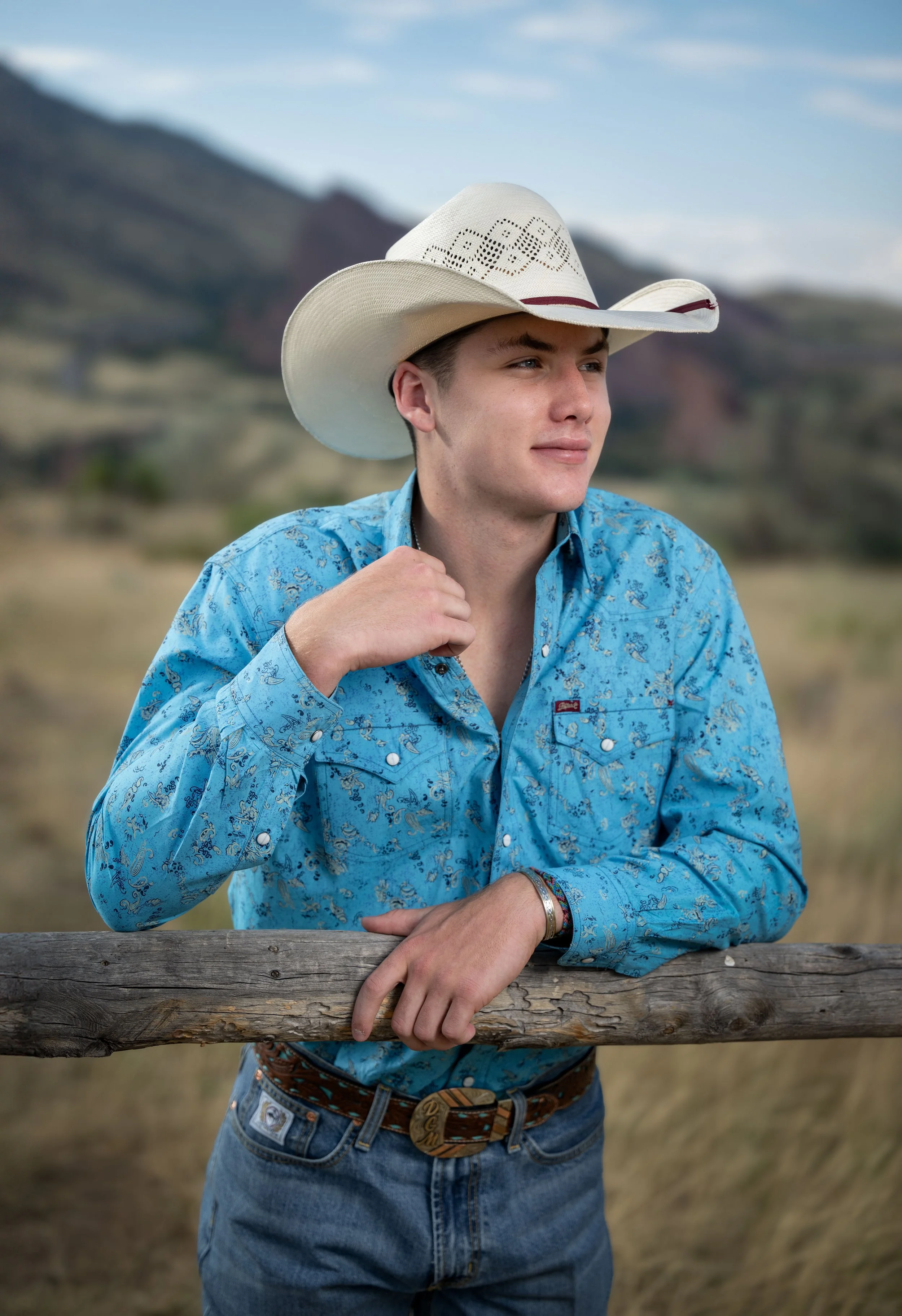 Young man in a cowboy hat and blue western shirt leaning on a wooden fence, outdoors with a mountainous landscape in the background.