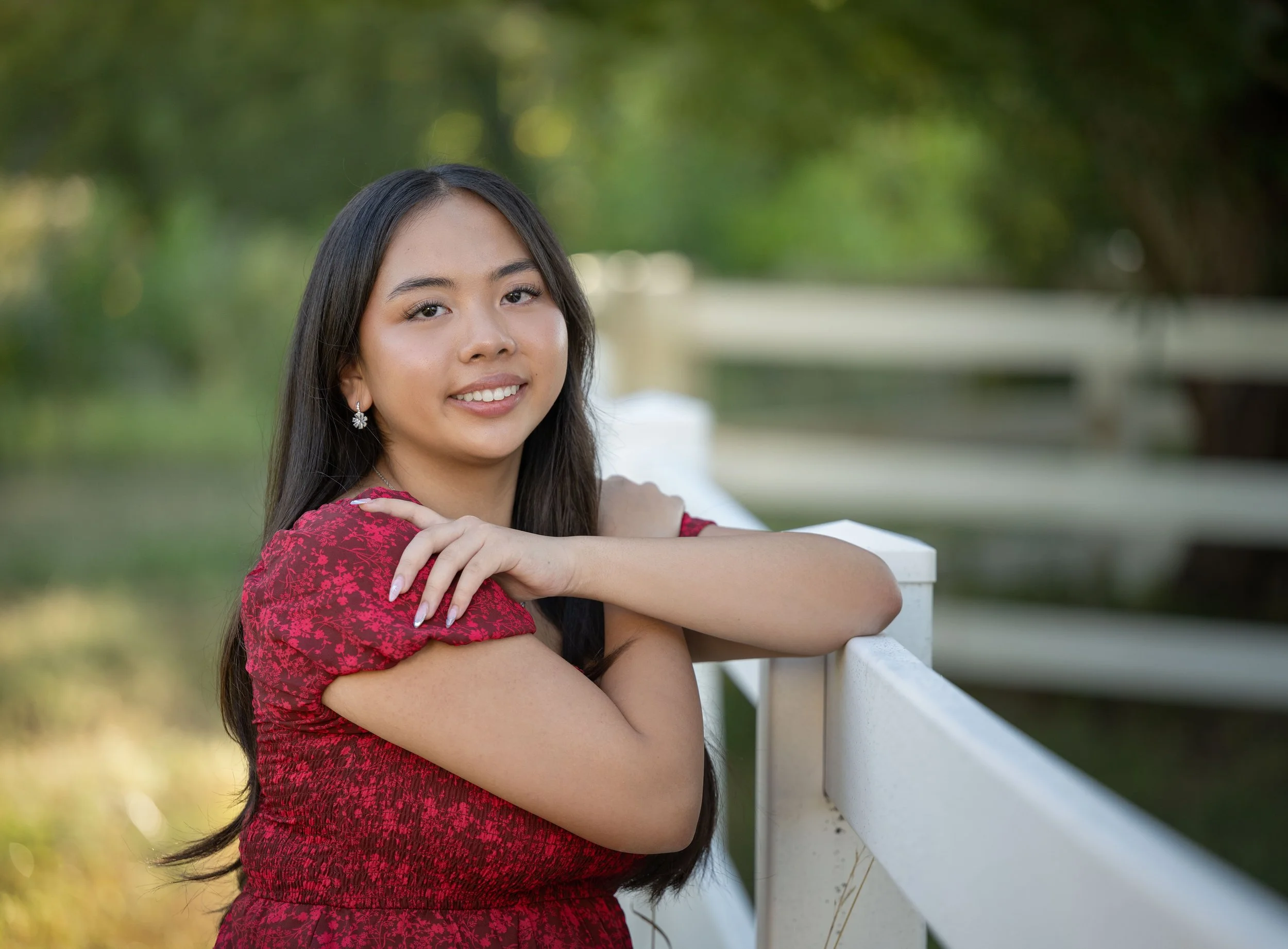 A young woman with long dark hair wearing a red dress and earrings, smiling and resting her arms on a white fence outdoors.